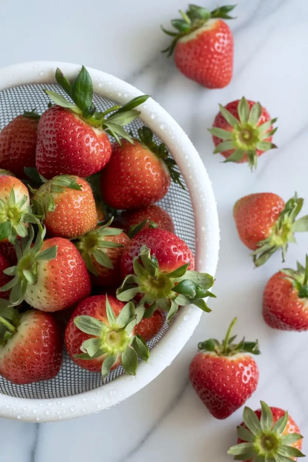 White colander holds fresh whole strawberries with green tops on a marble countertop. The strawberries look ripe and juicy, highlighting fresh fruit for homemade strawberry jam or freezer jam recipe.

