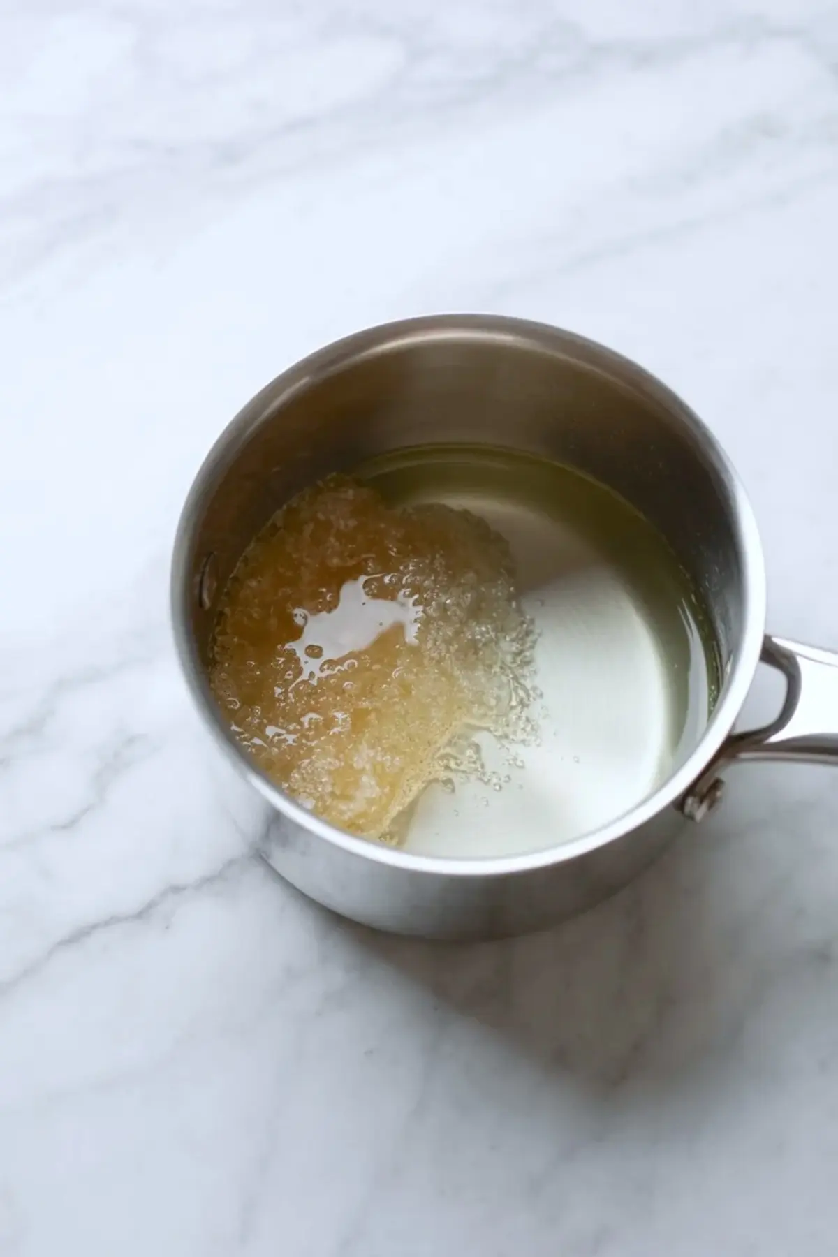 Small stainless steel saucepan holds blooming gelatin in water on a white marble surface. The gelatin forms a soft translucent mound in clear liquid, showing the early step of homemade strawberry freezer jam preparation.
