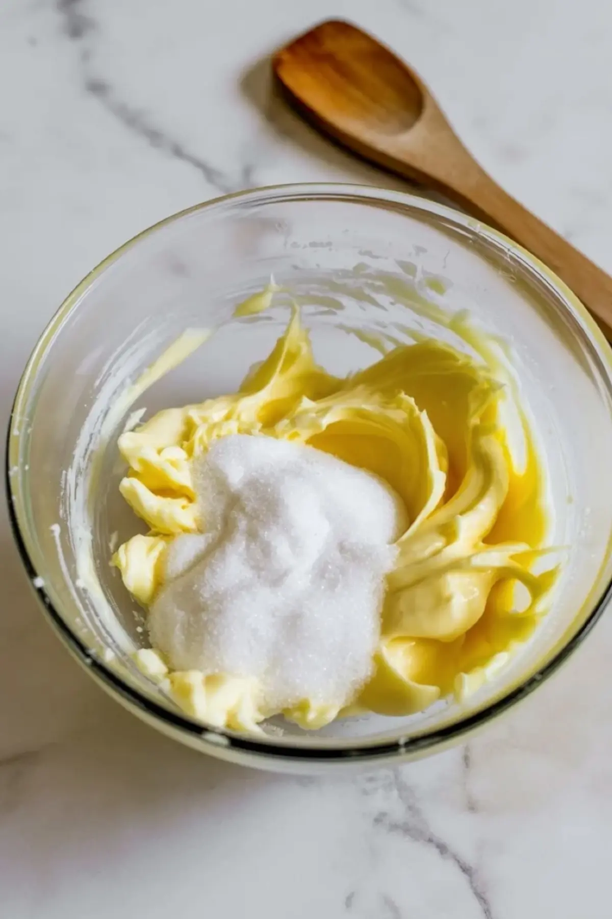Glass bowl holds softened butter and white granulated sugar, wooden spoon rests nearby on marble counter during cookie dough preparation.
