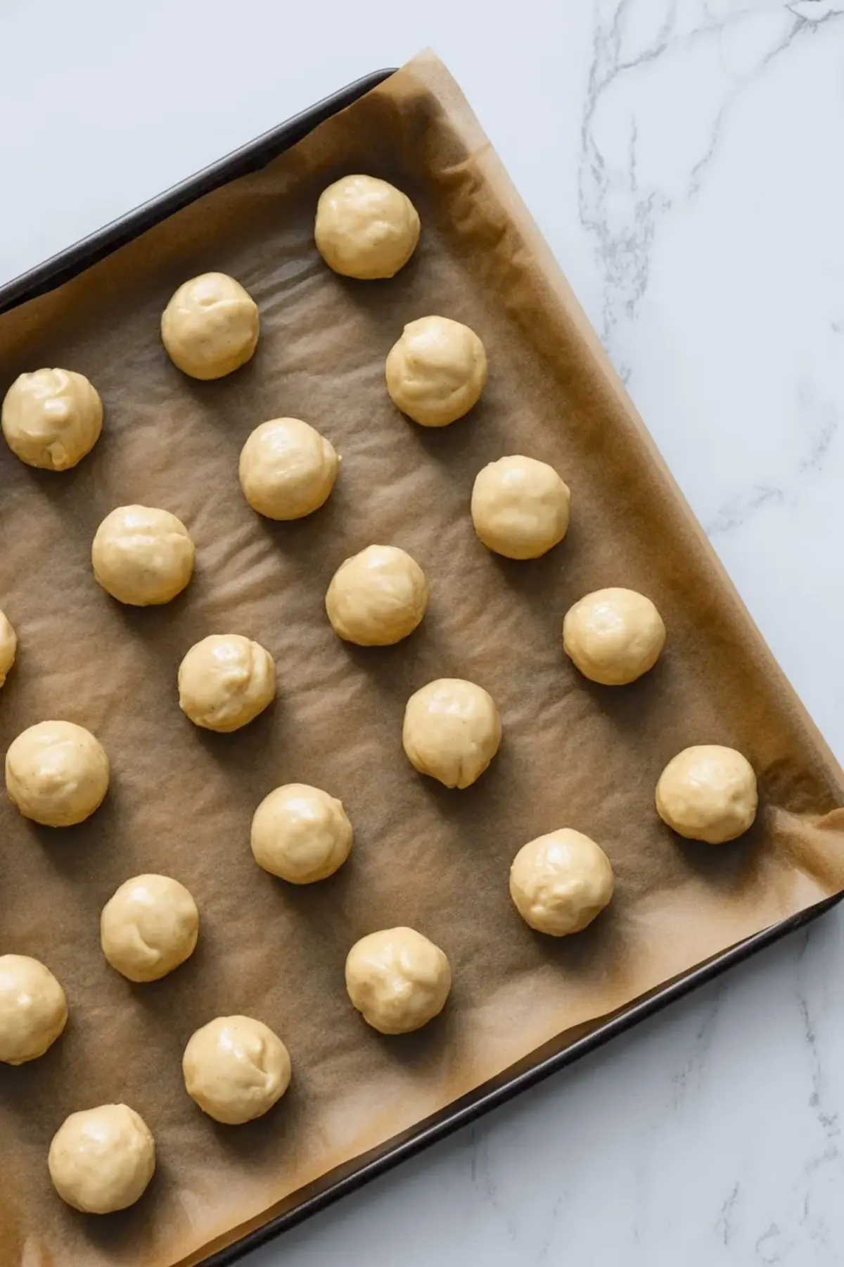 Round cookie dough balls sit spaced on a parchment lined baking sheet, arranged in neat rows on a marble surface, showing unbaked thumbprint cookies ready for the oven.
