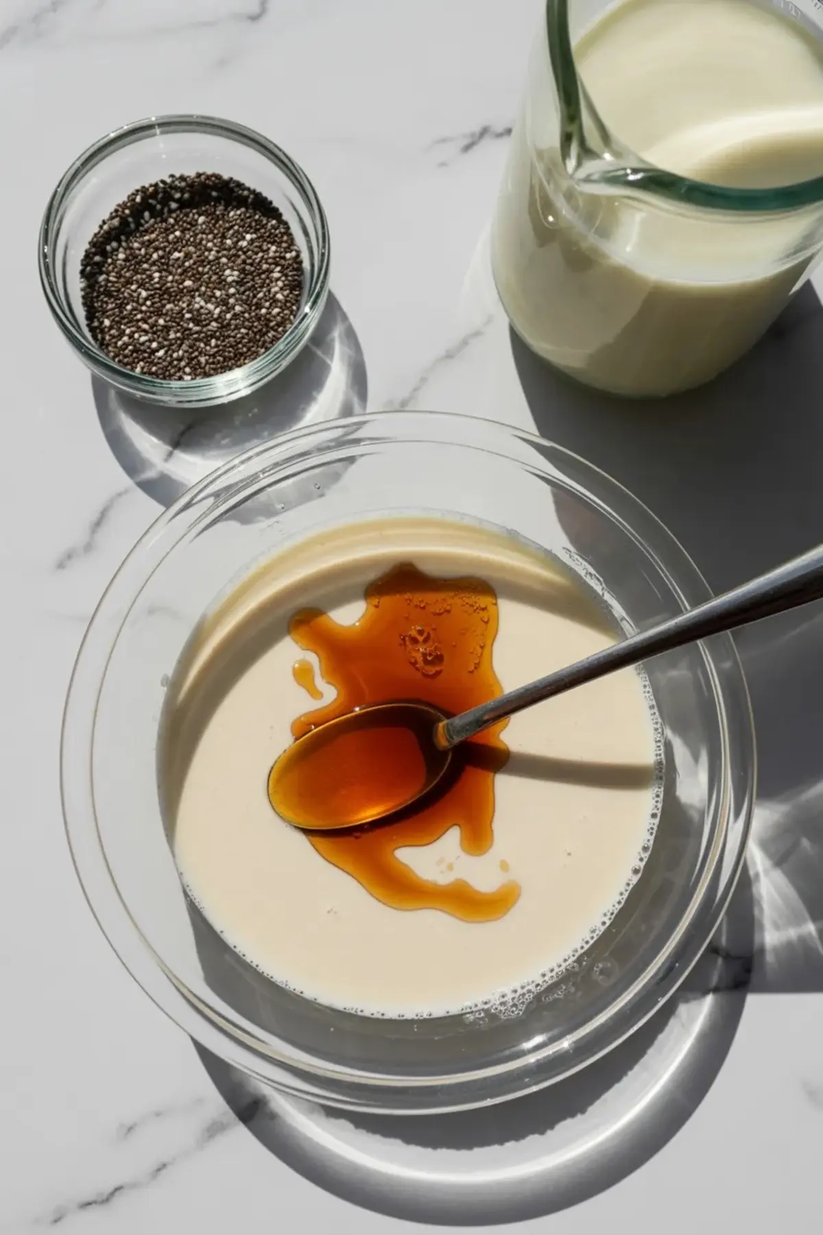 Clear glass bowl filled with creamy plant-based milk and maple syrup, with a spoon resting in the center, next to a small bowl of chia seeds and a glass jug of milk on a white marble surface.
