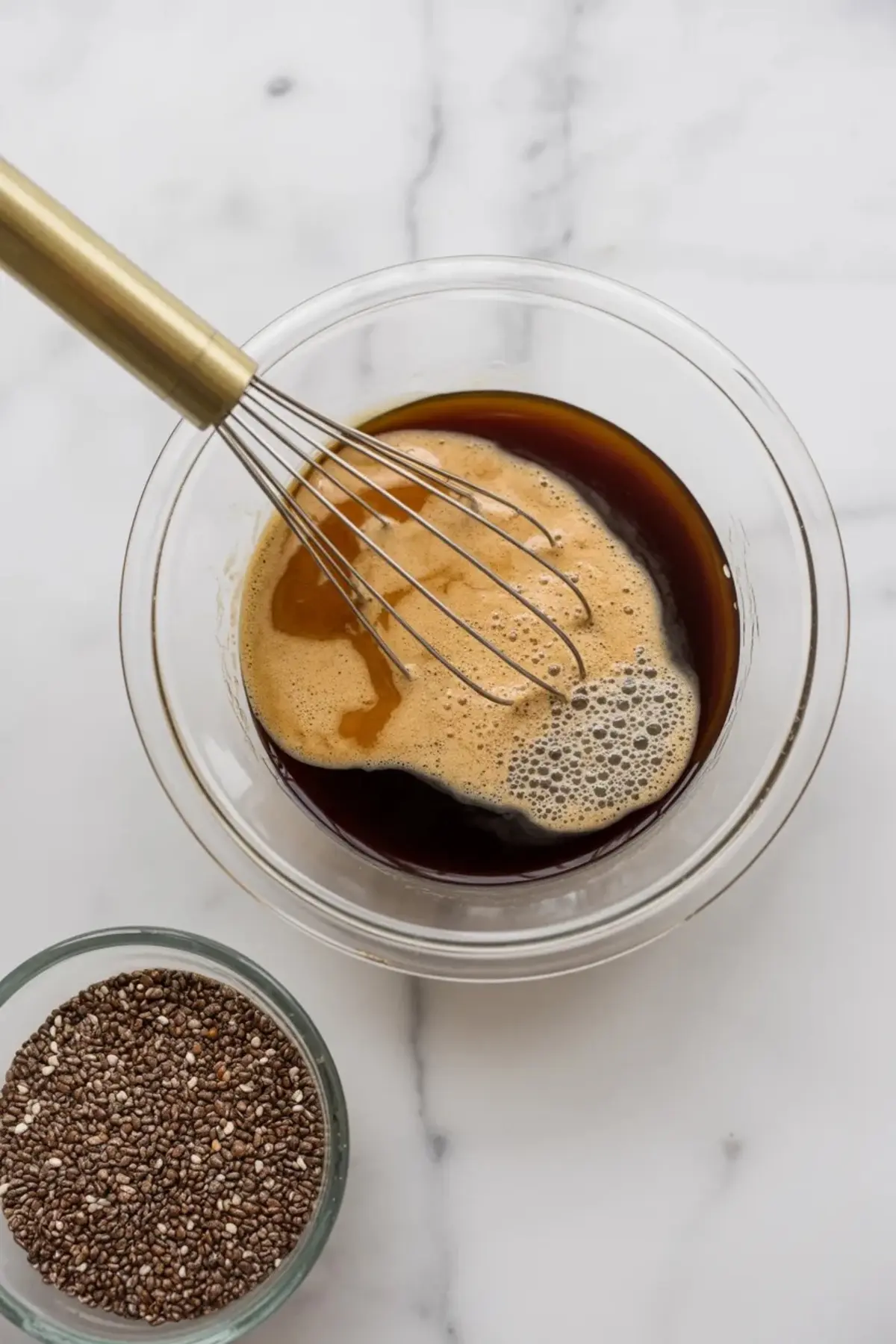 Glass bowl containing dark coffee mixture being whisked with a golden whisk, with visible foam and a small bowl of chia seeds placed nearby on a marble surface.
