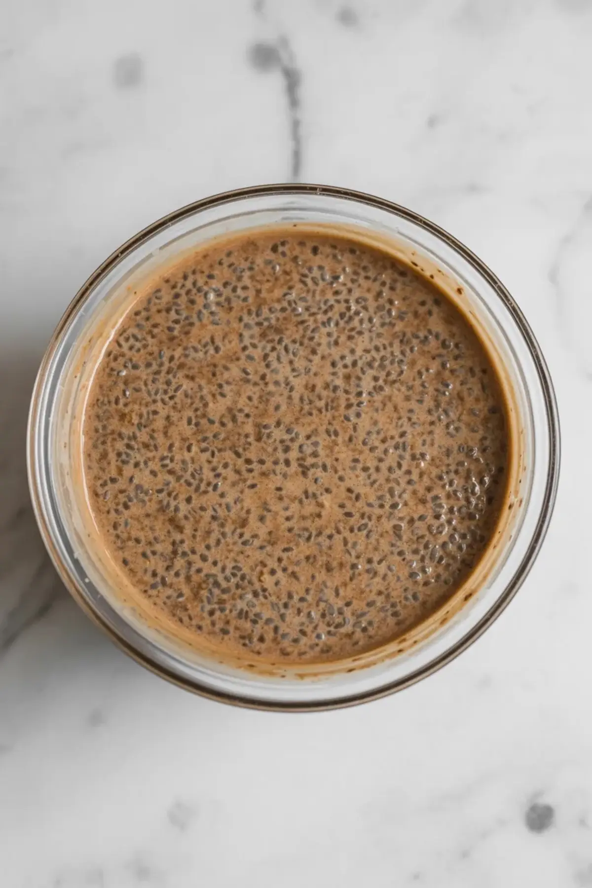 Thick chia pudding mixture in a glass bowl, featuring soaked chia seeds in a rich brown liquid, set on a clean white marble background.
