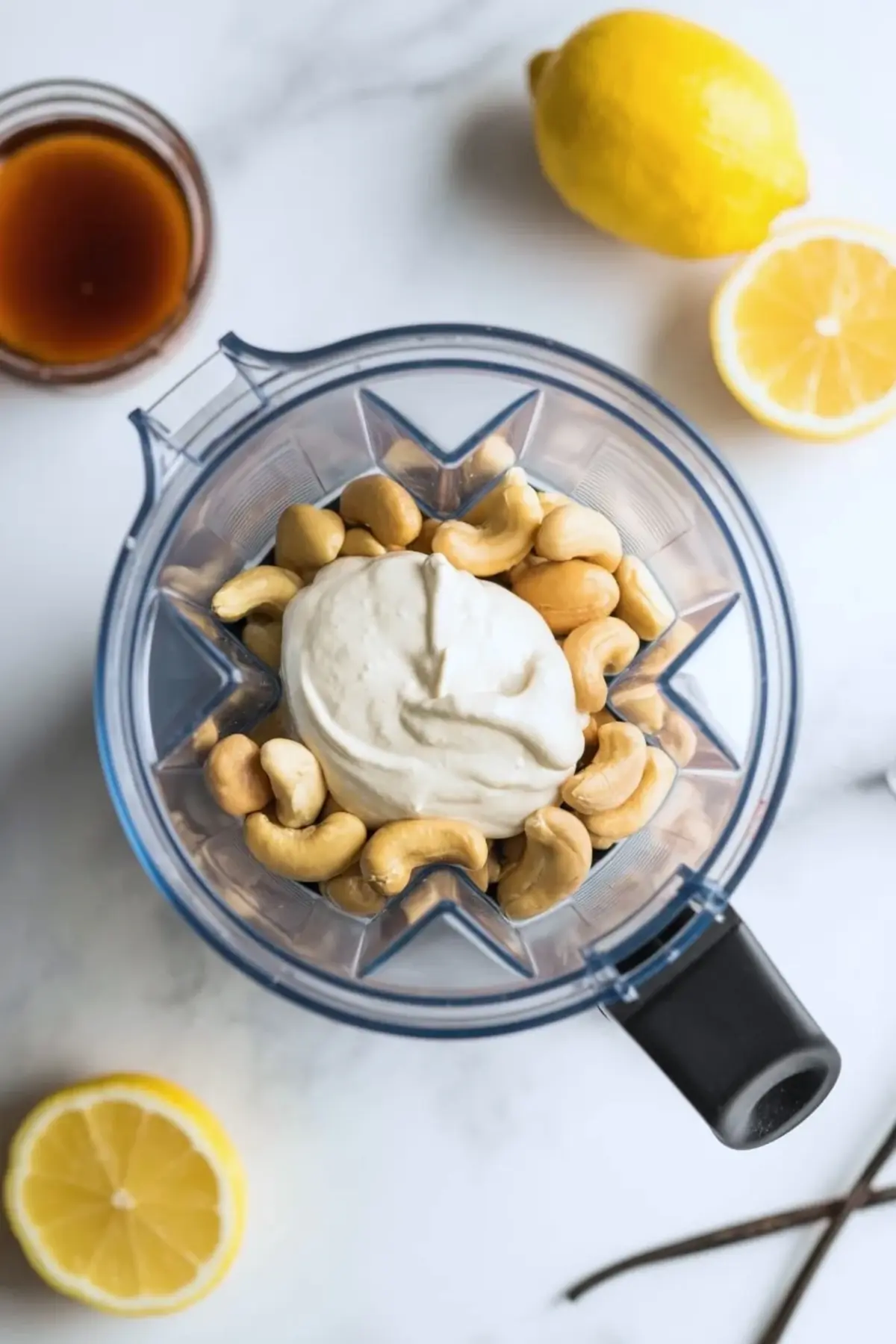 Blender filled with raw cashews and a creamy white mixture, surrounded by lemon halves, whole lemon, vanilla beans, and a jar of maple syrup on a white surface.

