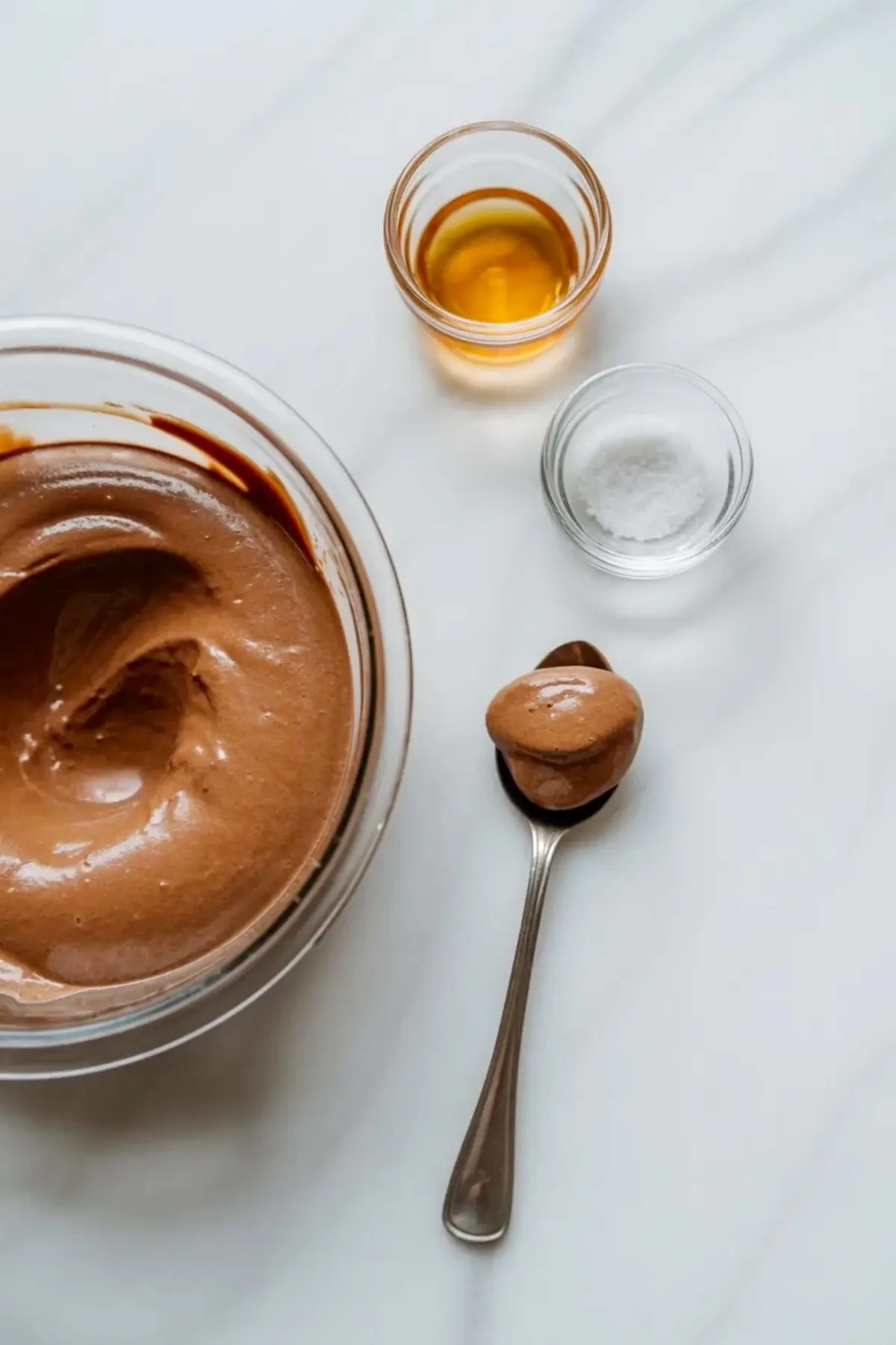 Glass mixing bowl holds thick chocolate mousse with a smooth swirl texture on a white marble surface. Small glass bowls with vanilla extract and salt sit nearby, and a spoon lifts a creamy scoop. This image shows vegan chocolate mousse preparation with simple ingredients for a dairy free dessert recipe.