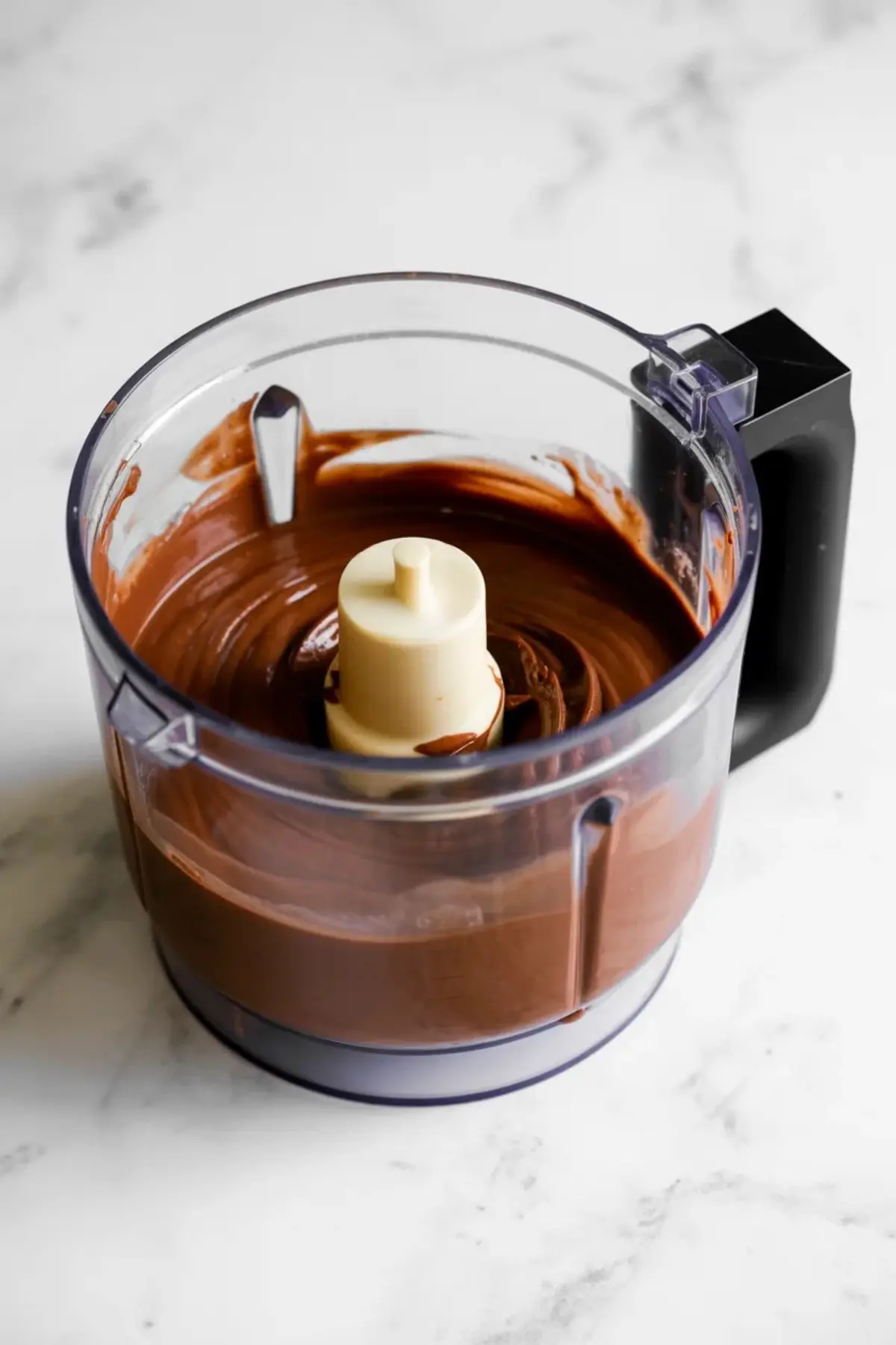 Food processor bowl holds blended chocolate tofu mixture with a smooth and thick consistency. The processor sits on a light marble surface. This image shows vegan chocolate mousse mixture being blended for a quick plant based dessert recipe.