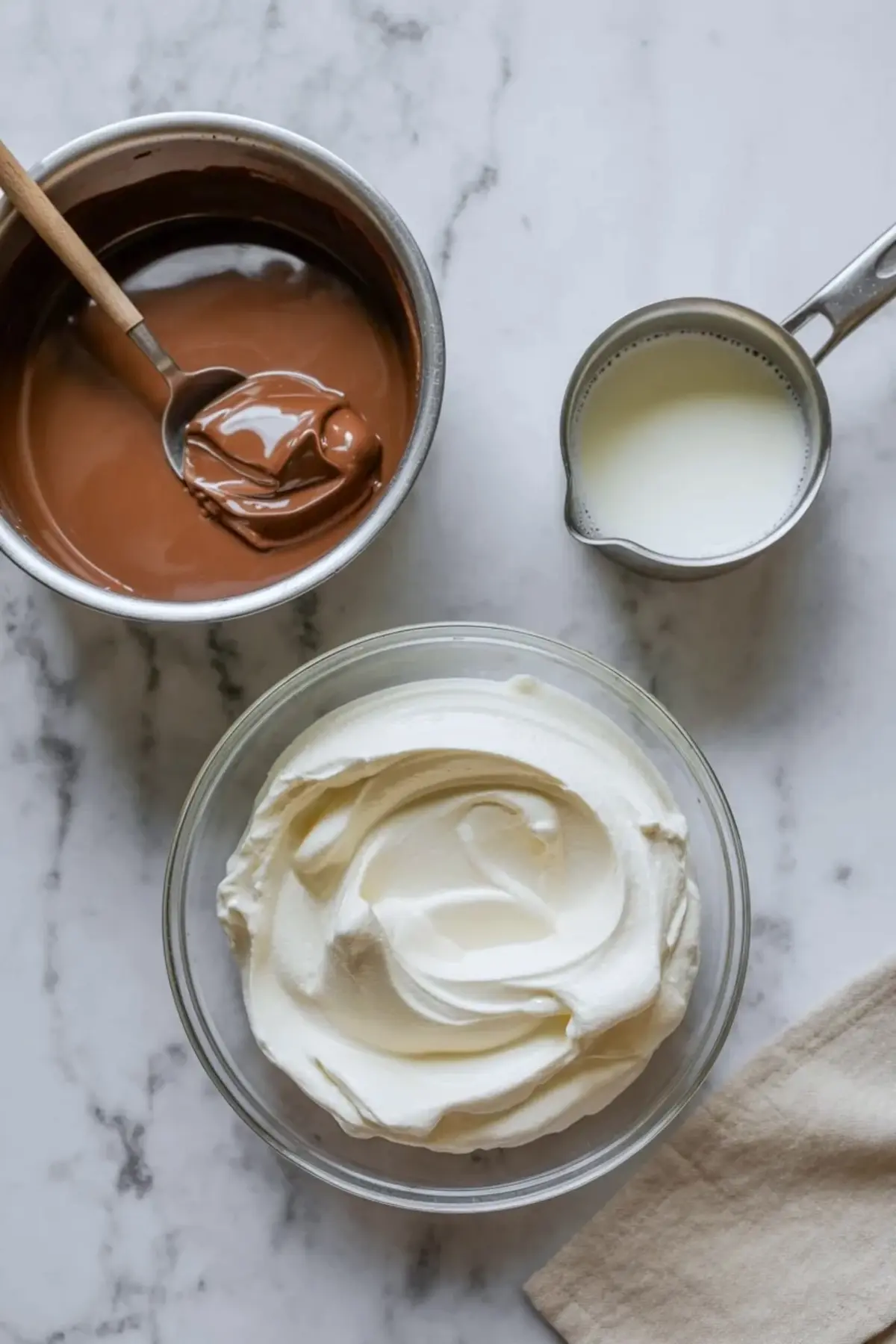 Overhead view of chocolate mousse ingredients with melted chocolate in a saucepan, heavy cream in a glass bowl, and milk in a metal measuring cup on marble.
