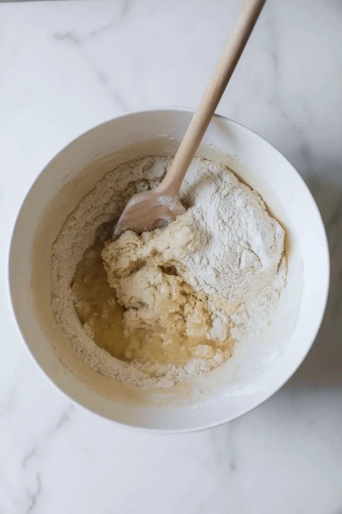 Mixing bowl holds flour, milk mixture, and butter while a wooden spoon combines the ingredients to form enriched sweet bread dough for tsoureki.
