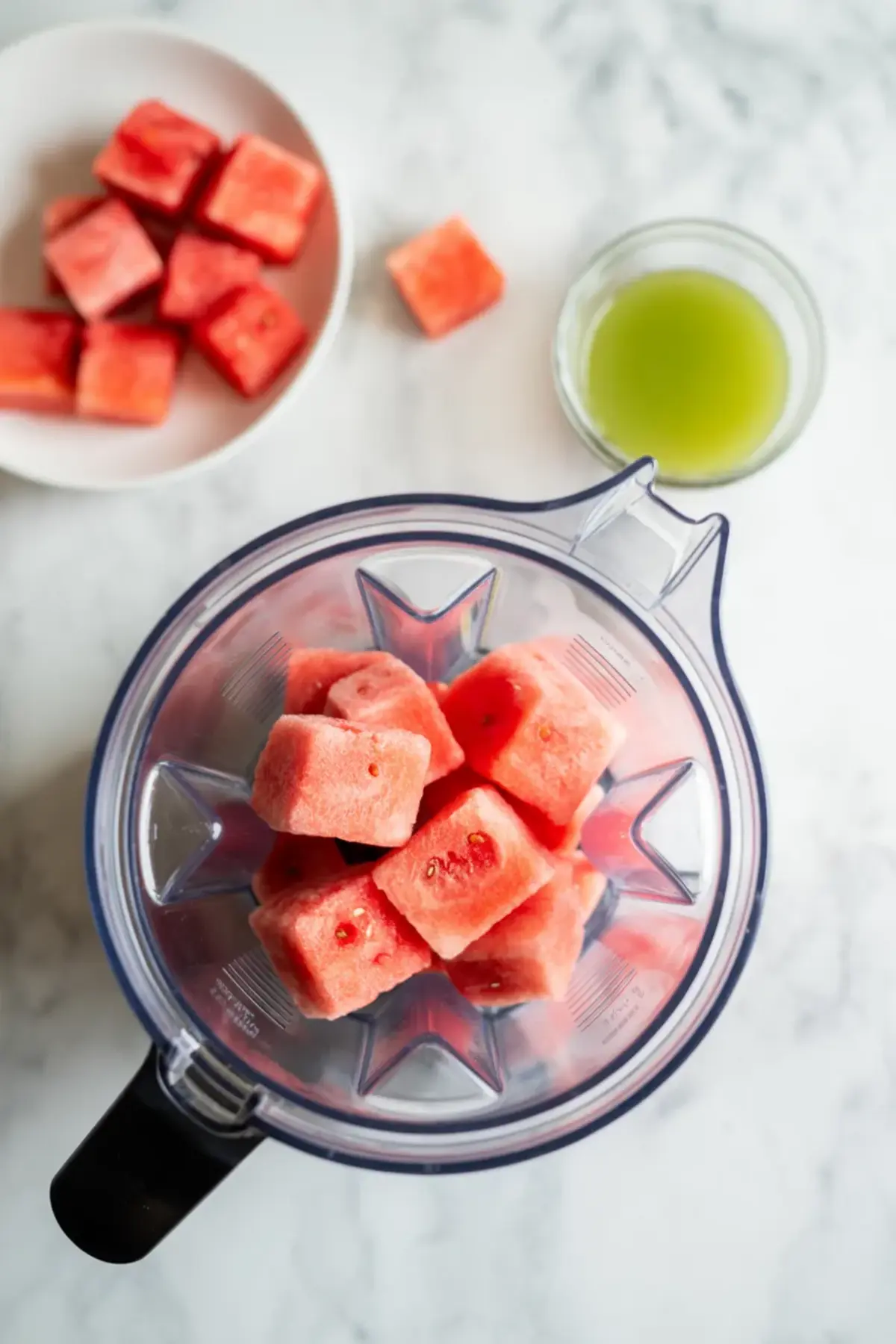 Top-down view of frozen watermelon cubes in a blender with a bowl of fresh watermelon chunks and a small glass bowl of lime juice nearby, showing simple ingredients for healthy fruit sorbet preparation.
