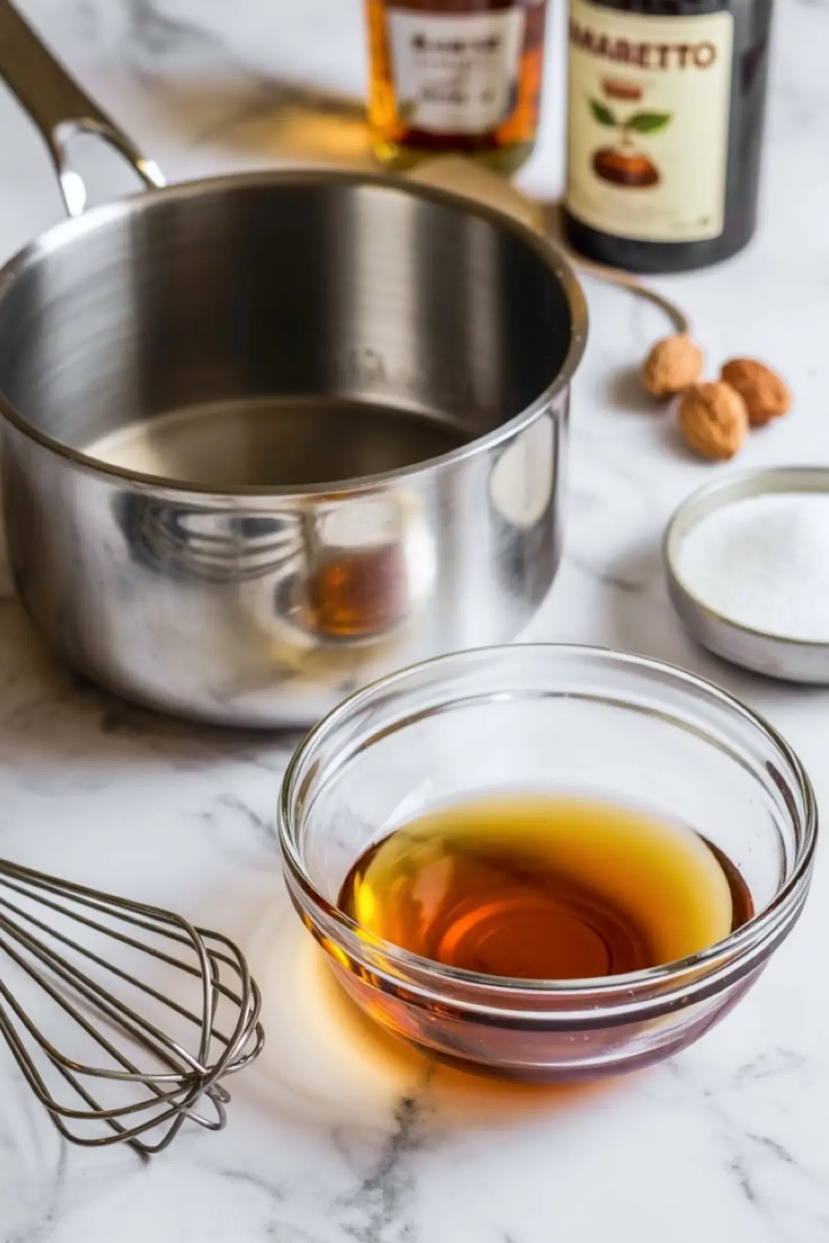Glass bowl holds amber amaretto liqueur beside a whisk, saucepan, sugar, whole almonds, and amaretto bottles, showing ingredients prepared for almond amaretto cake recipe.