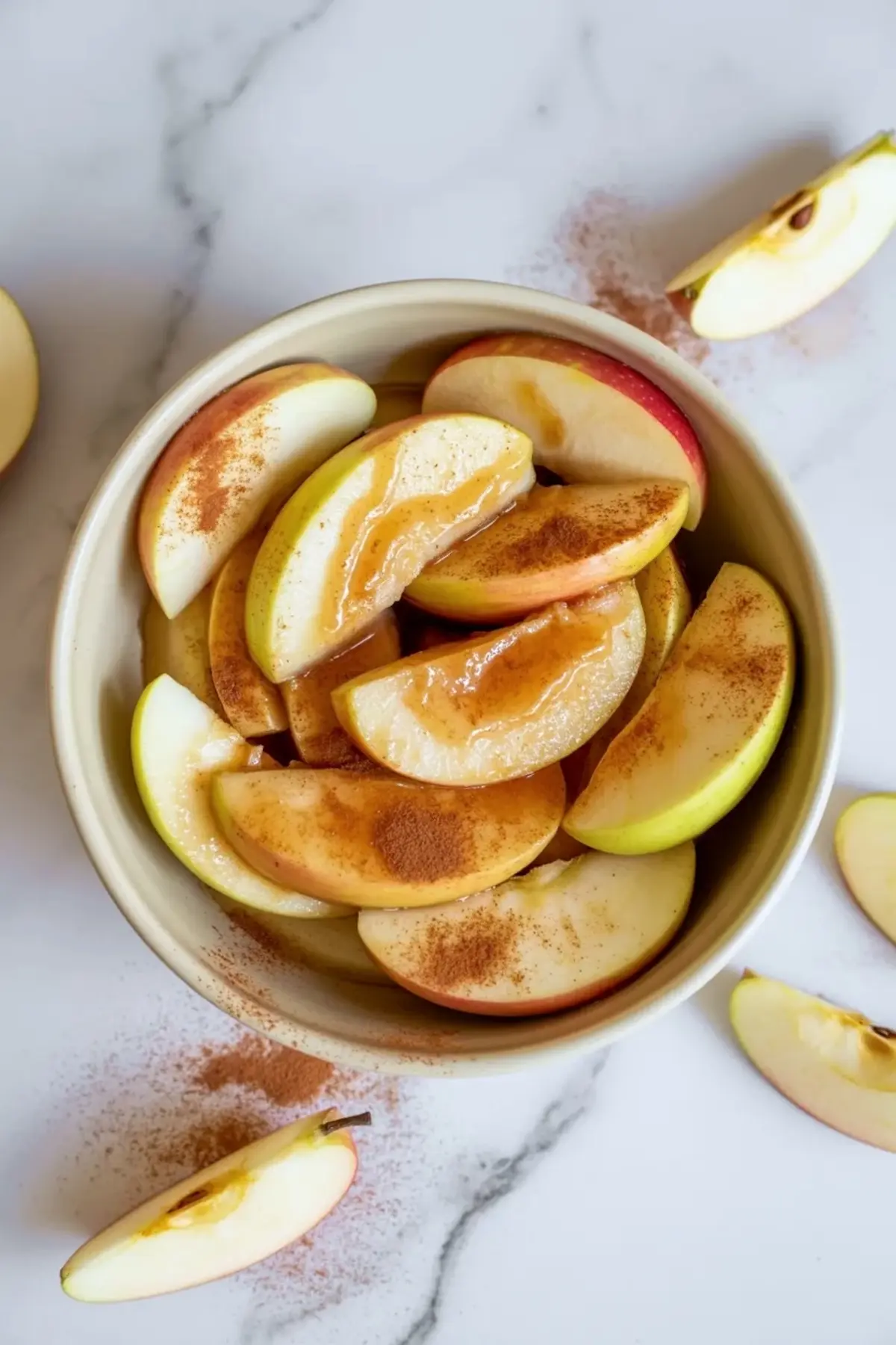Bowl of sliced apples for apple coffee cake dusted with cinnamon on a white marble counter.