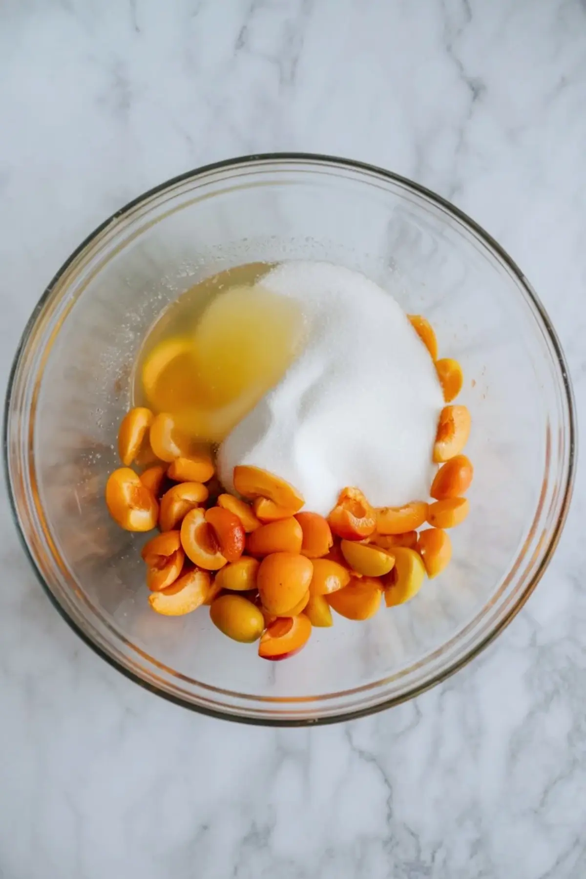 Apricot halves and sugar sit in a glass bowl on a marble surface before cooking apricot jam.