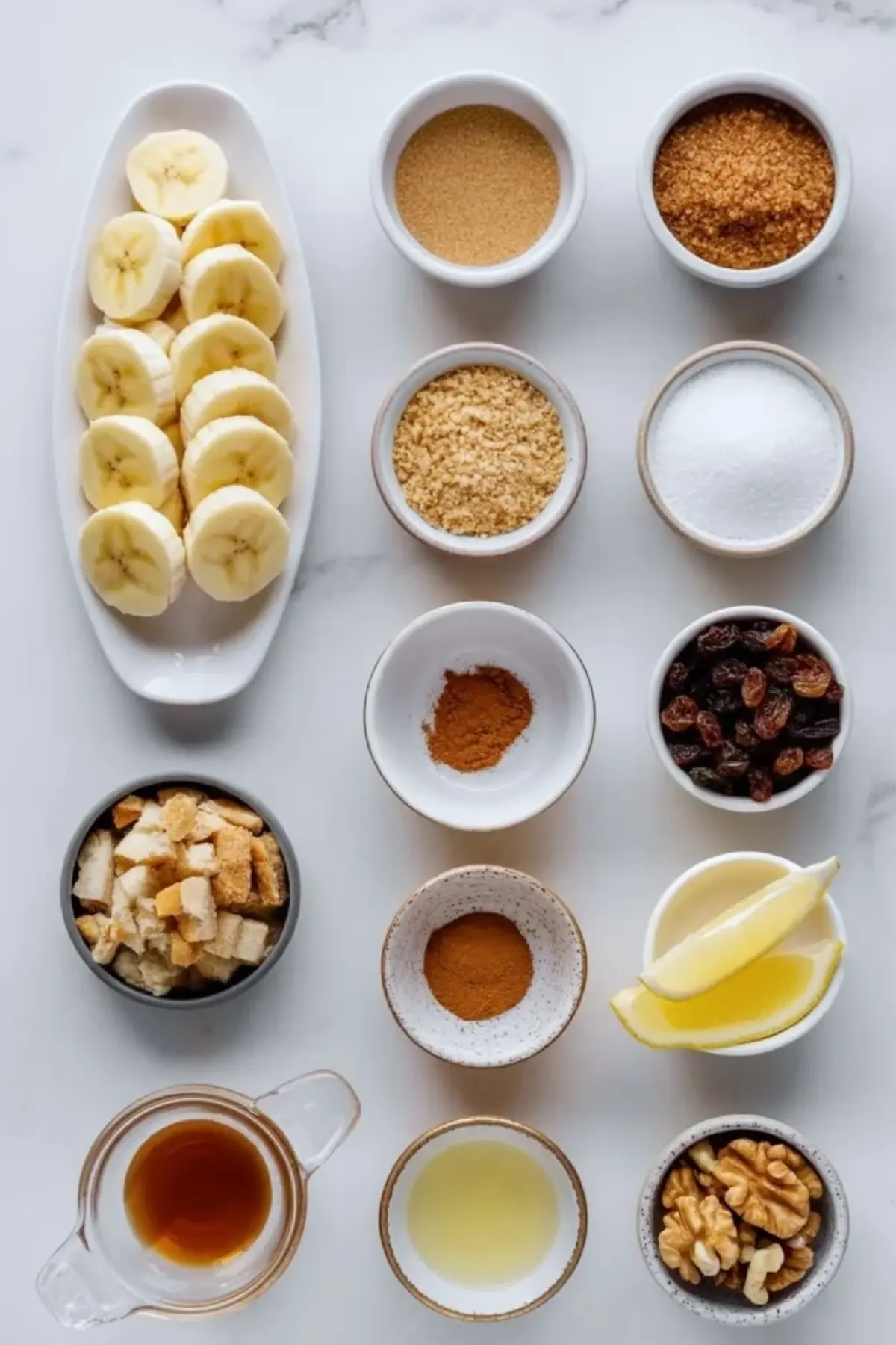 Ingredients for banana strudel arranged on white marble with sliced bananas, brown sugar, breadcrumbs, cinnamon, raisins, lemon, vanilla, and walnuts.