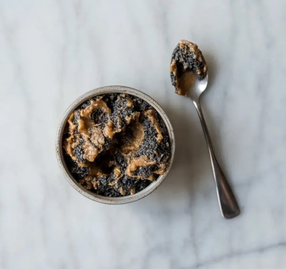 Small bowl contains dark black sesame paste or crumble with creamy swirls while a metal spoon holds a bite, set on a light marble surface for black sesame dessert filling inspiration.
