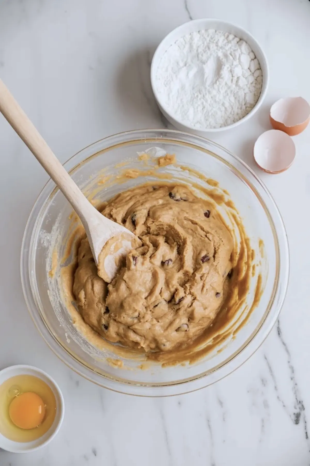 A glass mixing bowl holds thick cookie dough with chocolate chips while flour and eggs sit nearby, showing homemade cookie dough preparation on a marble countertop.
