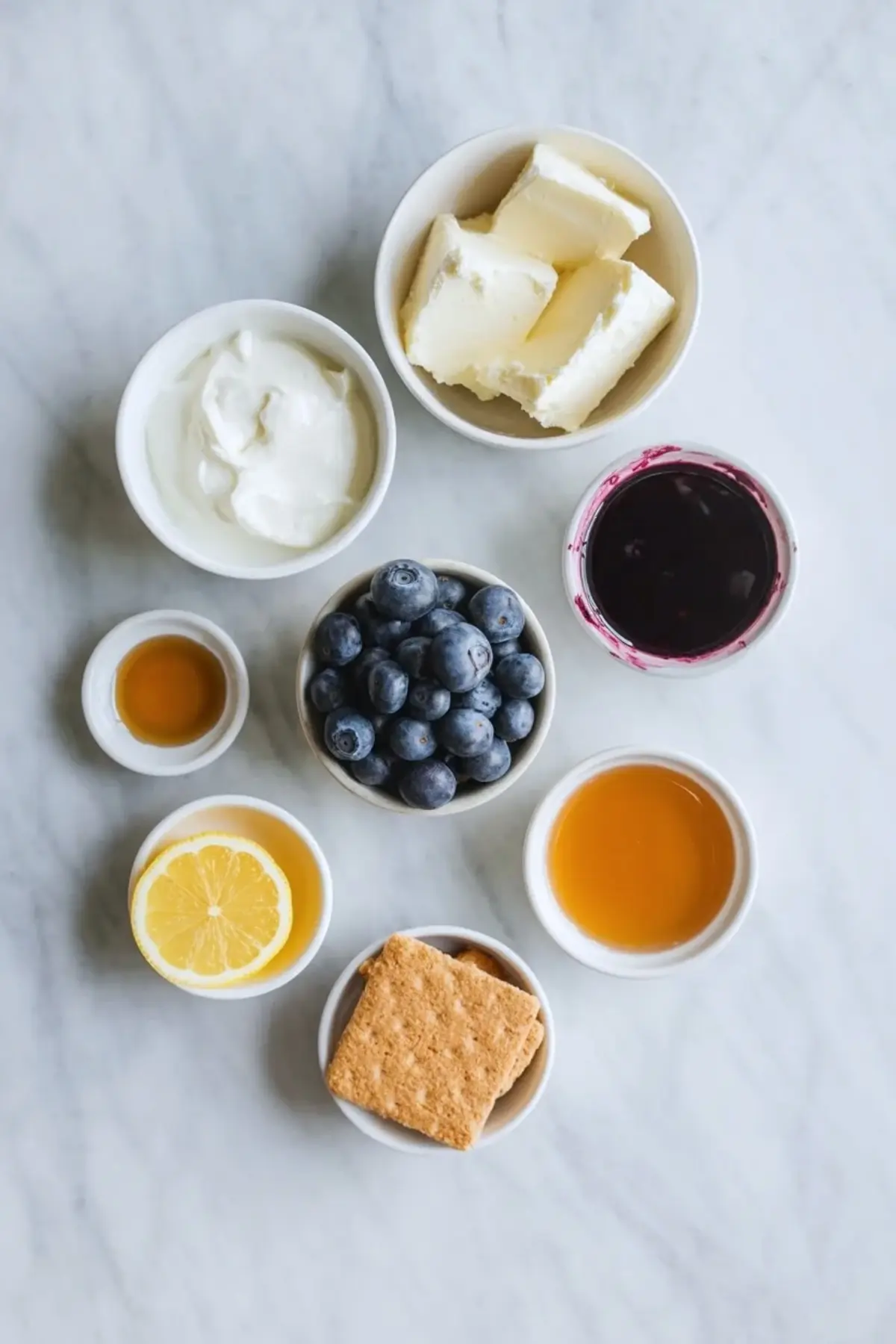 Ingredients for blueberry cheesecake yogurt bark arranged in small bowls, including blueberries, yogurt, cream cheese, lemon, honey, and graham crackers.