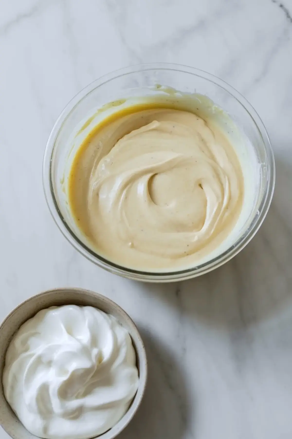 Blueberry vanilla cheesecake icebox cake filling in a glass bowl beside whipped cream, showing the smooth vanilla cheesecake layer before assembly on white marble.