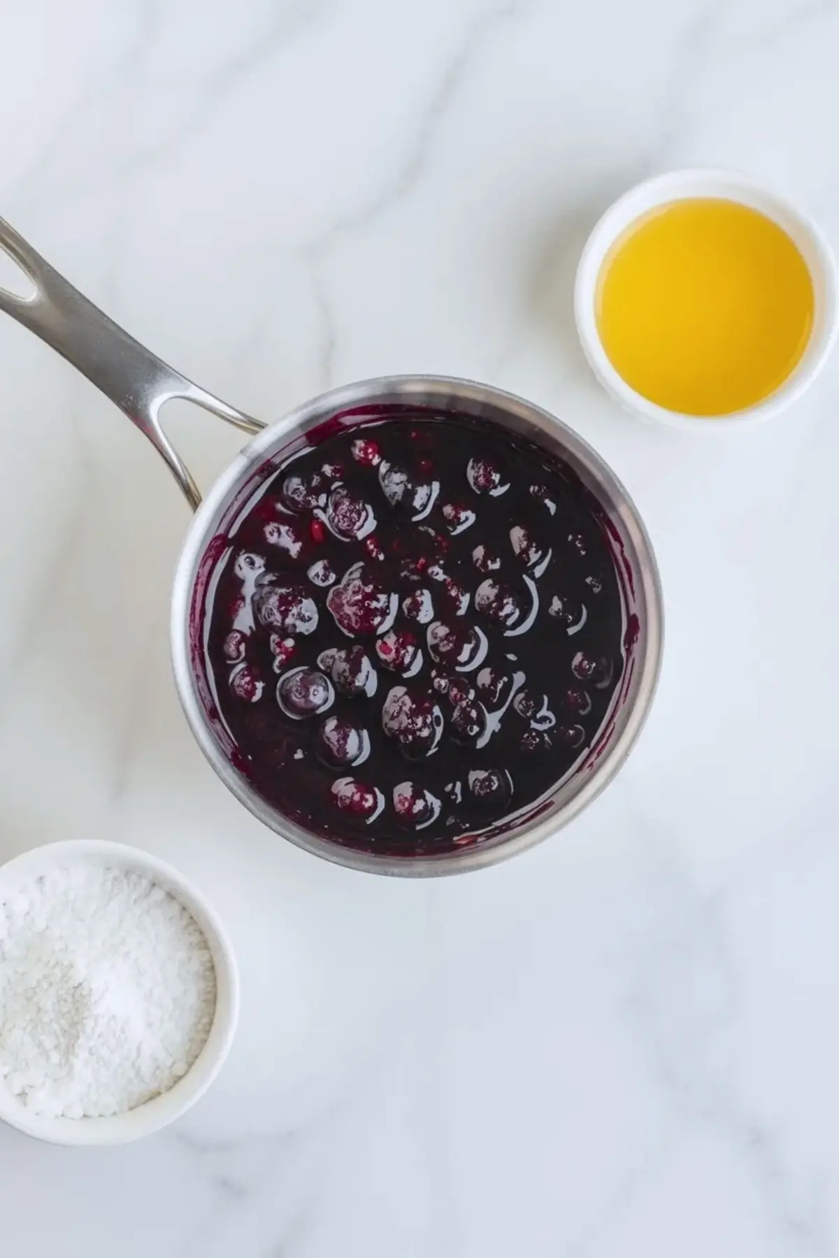 Homemade blueberry topping for blueberry vanilla cheesecake icebox cake simmering in a saucepan with cornstarch and lemon juice on white marble.