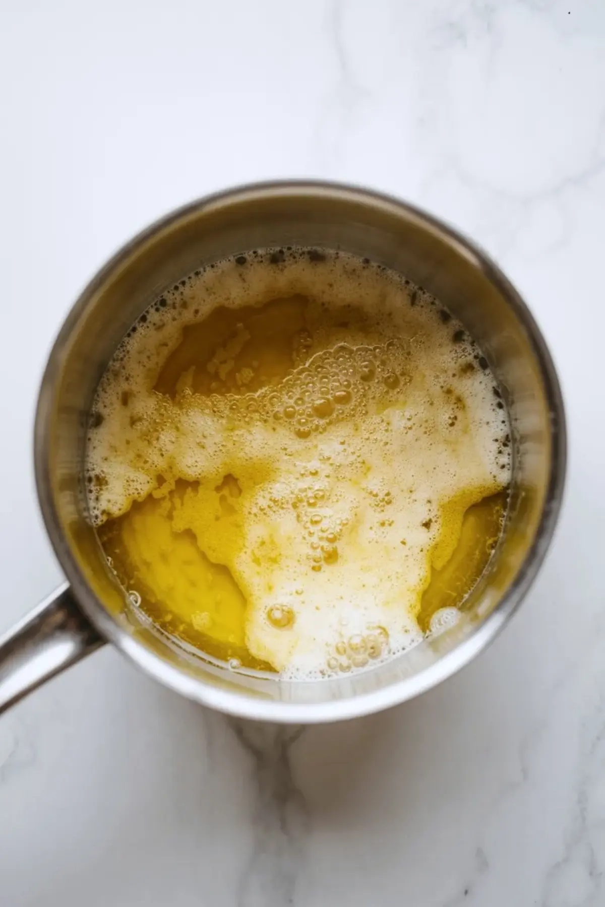 Top view shows browned butter with golden foam bubbling in a small stainless steel saucepan on a light marble countertop, step for brown butter baking ingredient.
