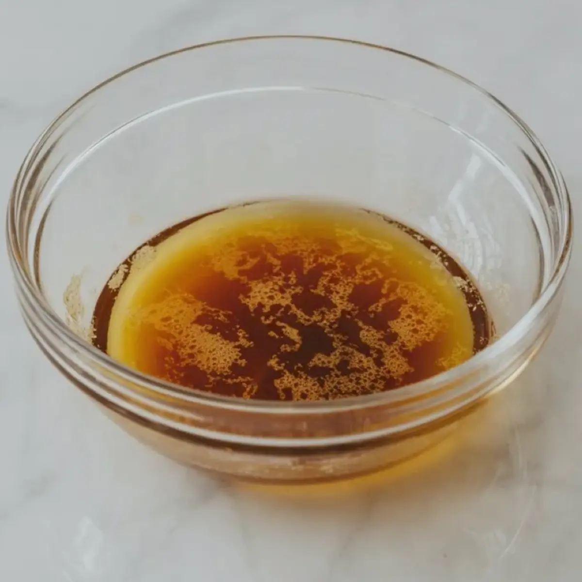 Clear glass bowl holds warm brown butter with deep amber color and small toasted milk solids on a light marble surface for brown butter frosting recipe.

