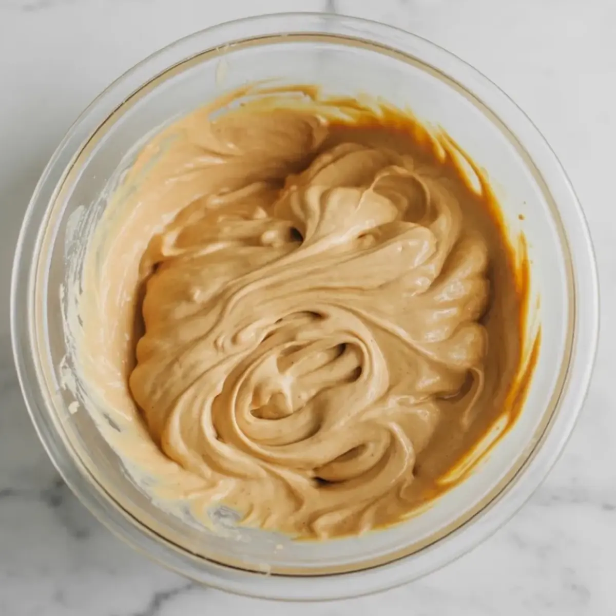 Glass mixing bowl contains smooth tan brown butter cream mixture with thick swirls from a spatula, step for brown butter cream cheese frosting.
