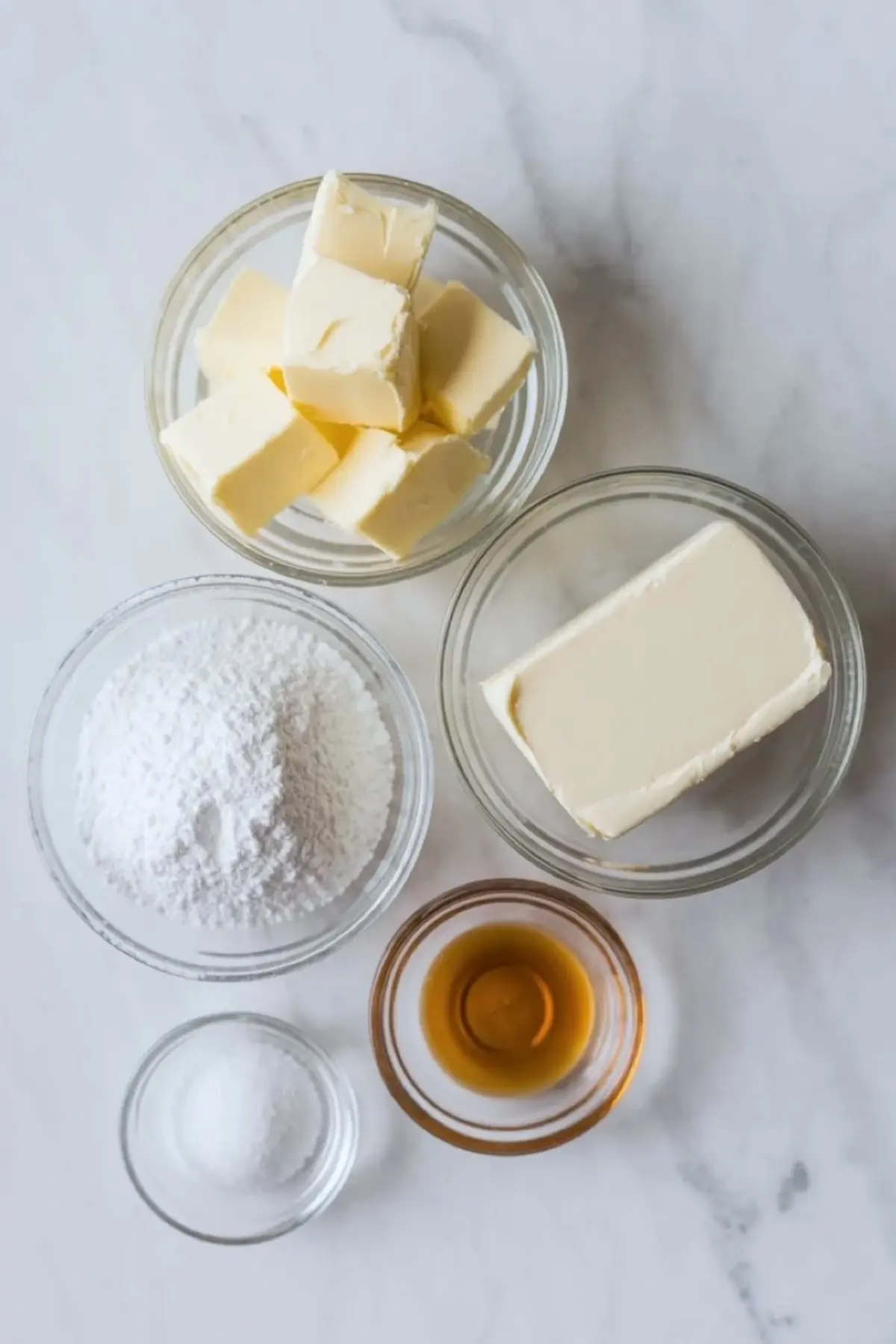 Flat lay shows baking ingredients in glass bowls with cubed butter, cream cheese block, powdered sugar, vanilla extract, and salt on a white marble countertop for cream cheese frosting recipe.
