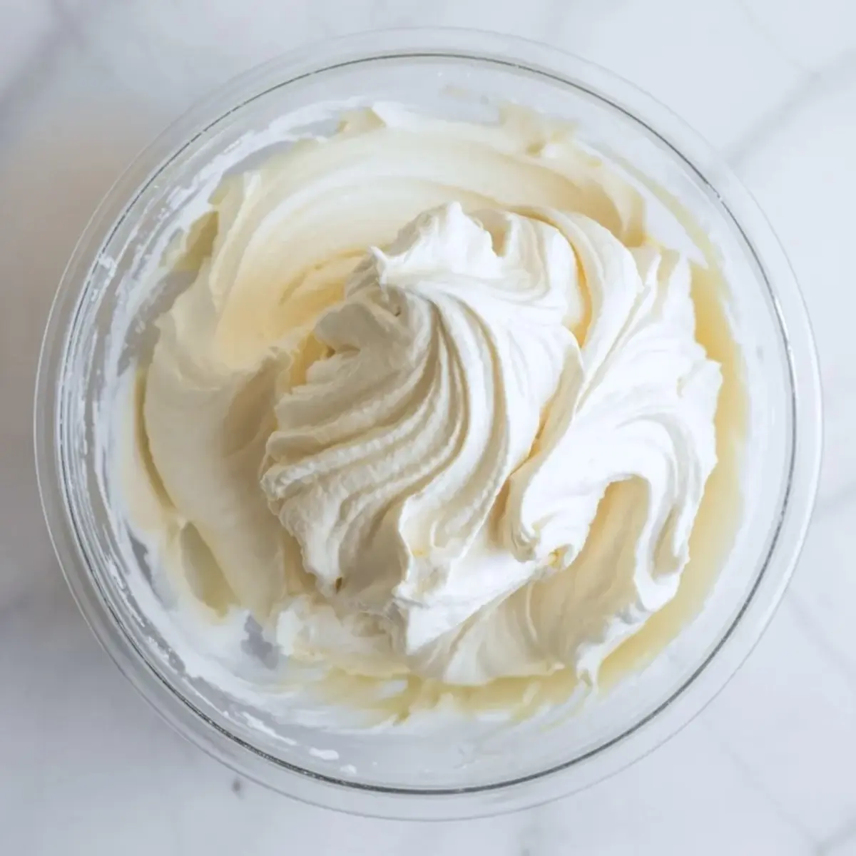 Glass bowl holds whipped cream cheese mixture with soft peaks and smooth texture on a marble surface for homemade cream cheese frosting base.
