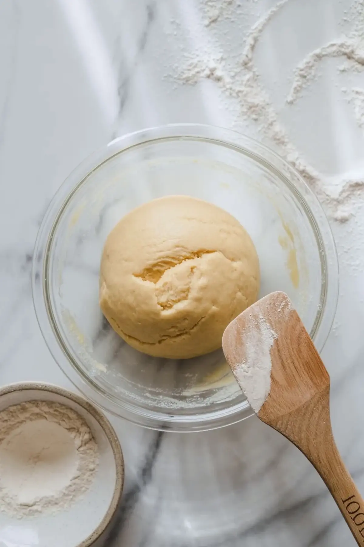 Glass bowl holds a smooth ball of sugar cookie dough while a wooden spatula and flour sit nearby on a marble countertop during cookie dough preparation.
