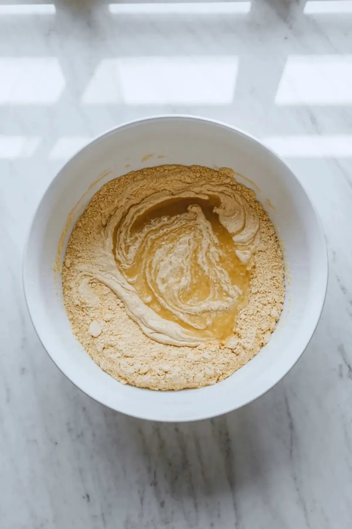 White mixing bowl holds cornmeal batter with melted butter or oil swirling into the mixture on a light marble countertop during buttermilk cornbread recipe preparation.

