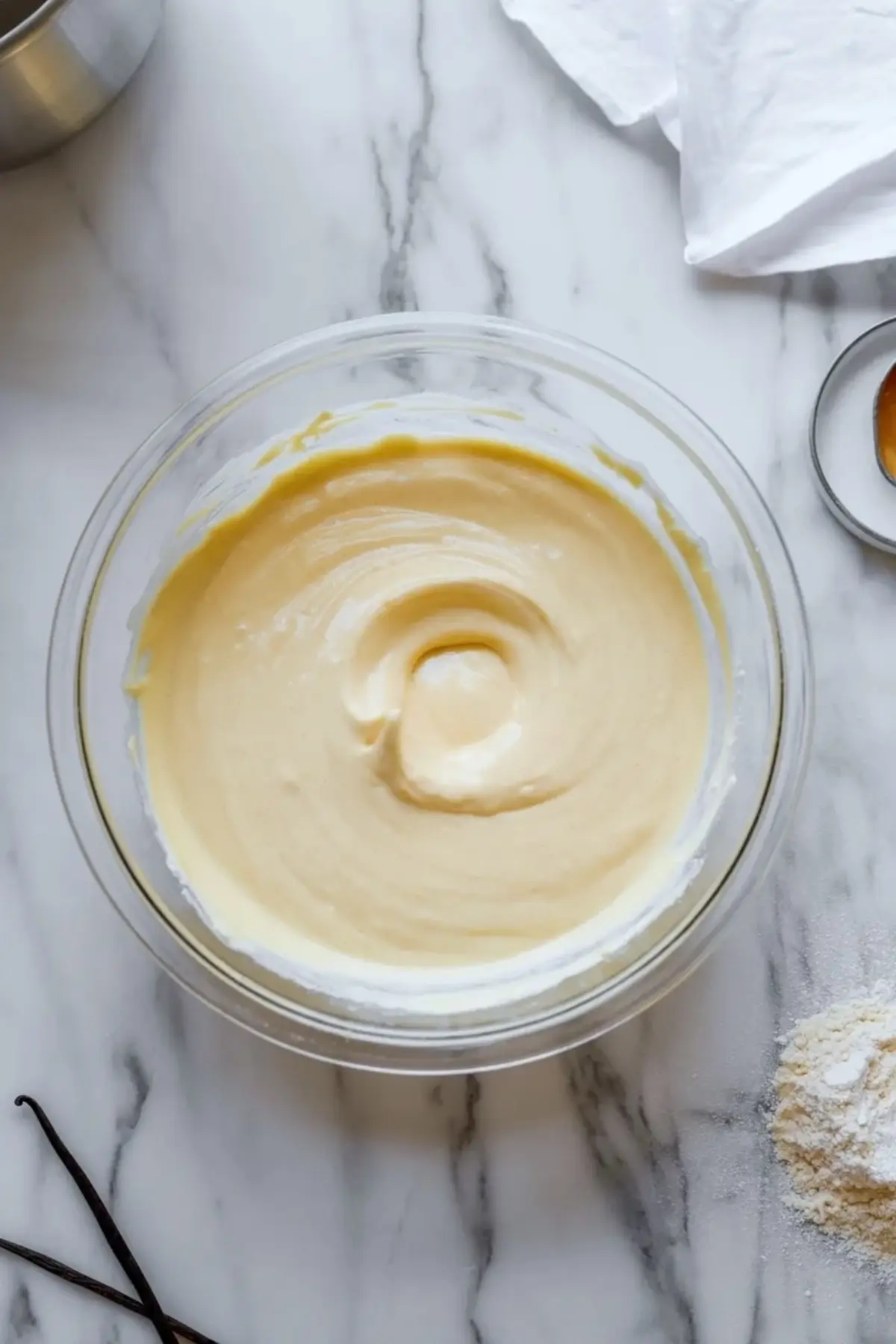 Glass bowl contains smooth creamy cheesecake batter with vanilla bean pods and flour on marble countertop during dessert filling preparation.
