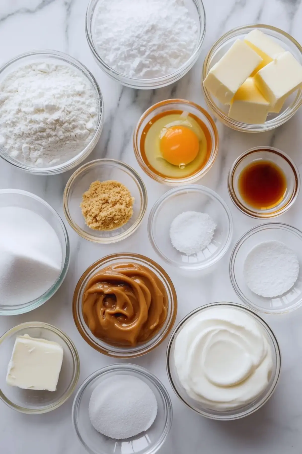 Small glass bowls display baking ingredients including flour, butter cubes, egg, vanilla extract, sugar, brown sugar, cream cheese, and caramel for caramel cheesecake cookies recipe.
