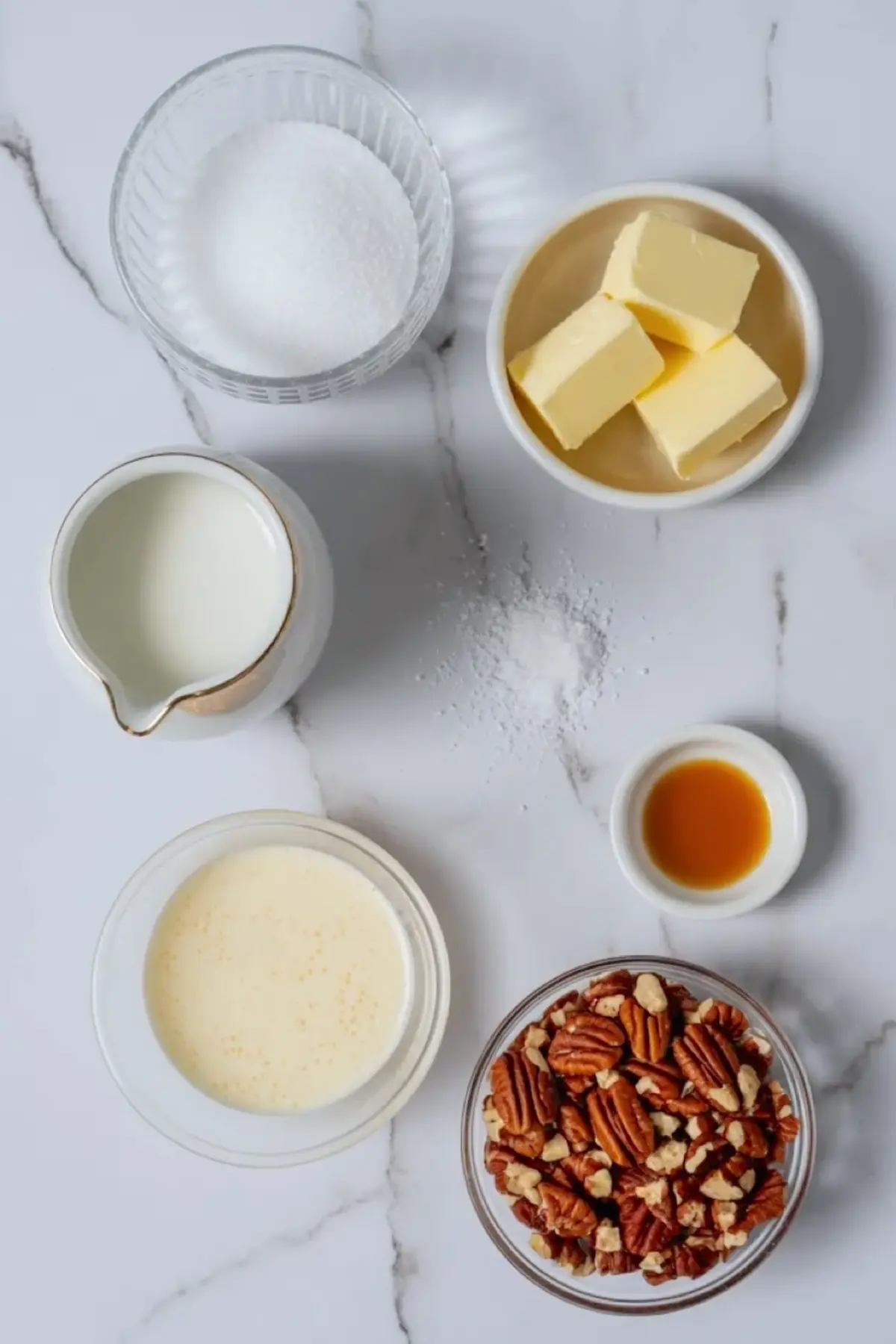 Ingredients for caramel pecan cake filling arranged with sugar, butter, cream, vanilla, and chopped pecans on white marble.