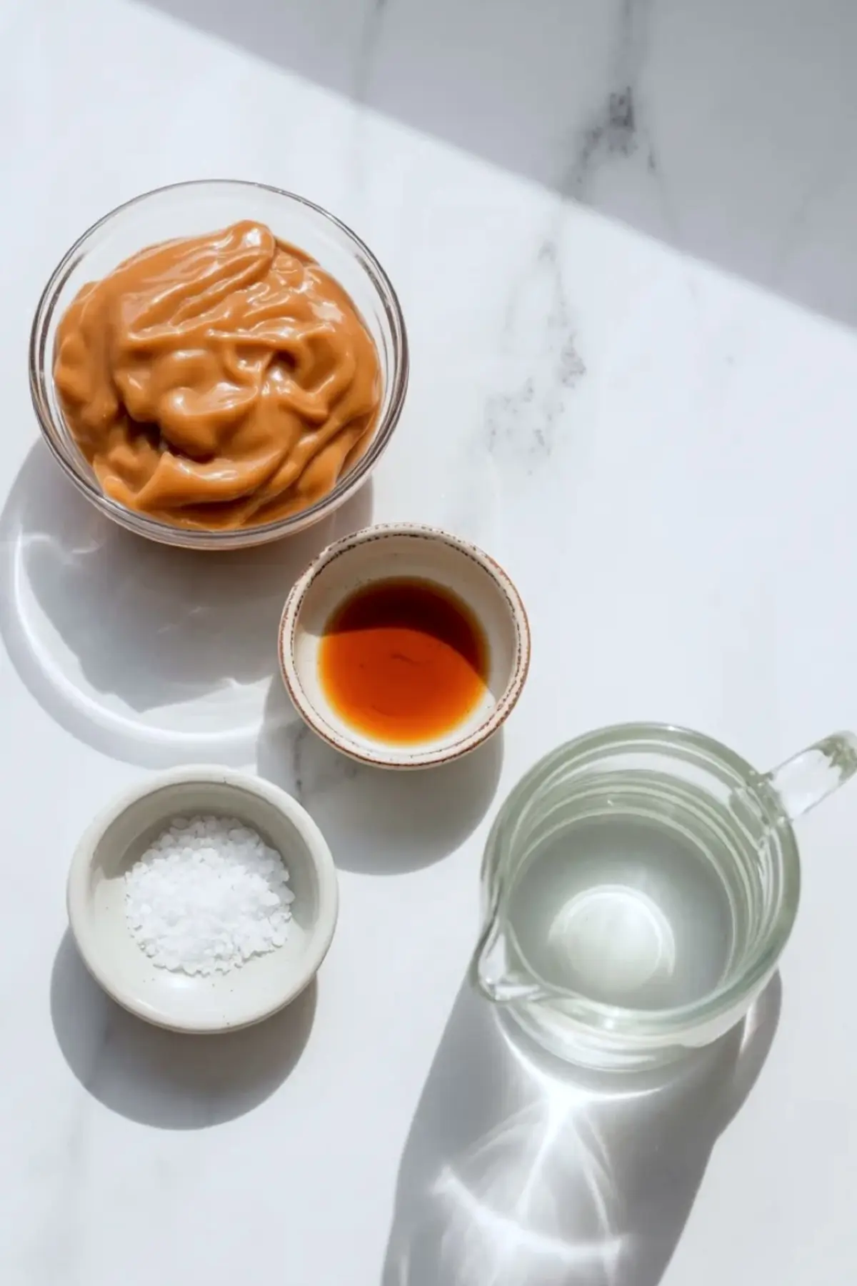 A bowl of thick caramel sauce sits on a white surface beside small bowls of vanilla extract and sea salt, and a glass measuring cup filled with water. The ingredients appear arranged for a caramel sauce recipe. This image shows simple ingredients used to make condensed milk caramel at home.