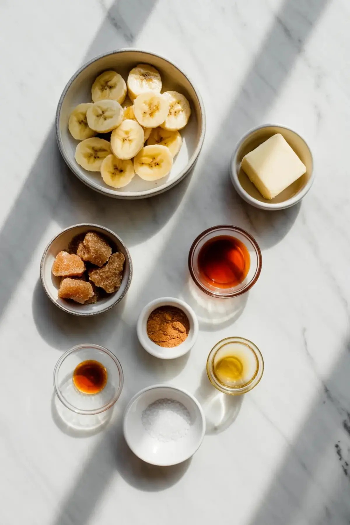 Ingredients for caramelized banana recipe arranged in small bowls with sliced bananas, butter, brown sugar, cinnamon, vanilla, and salt.