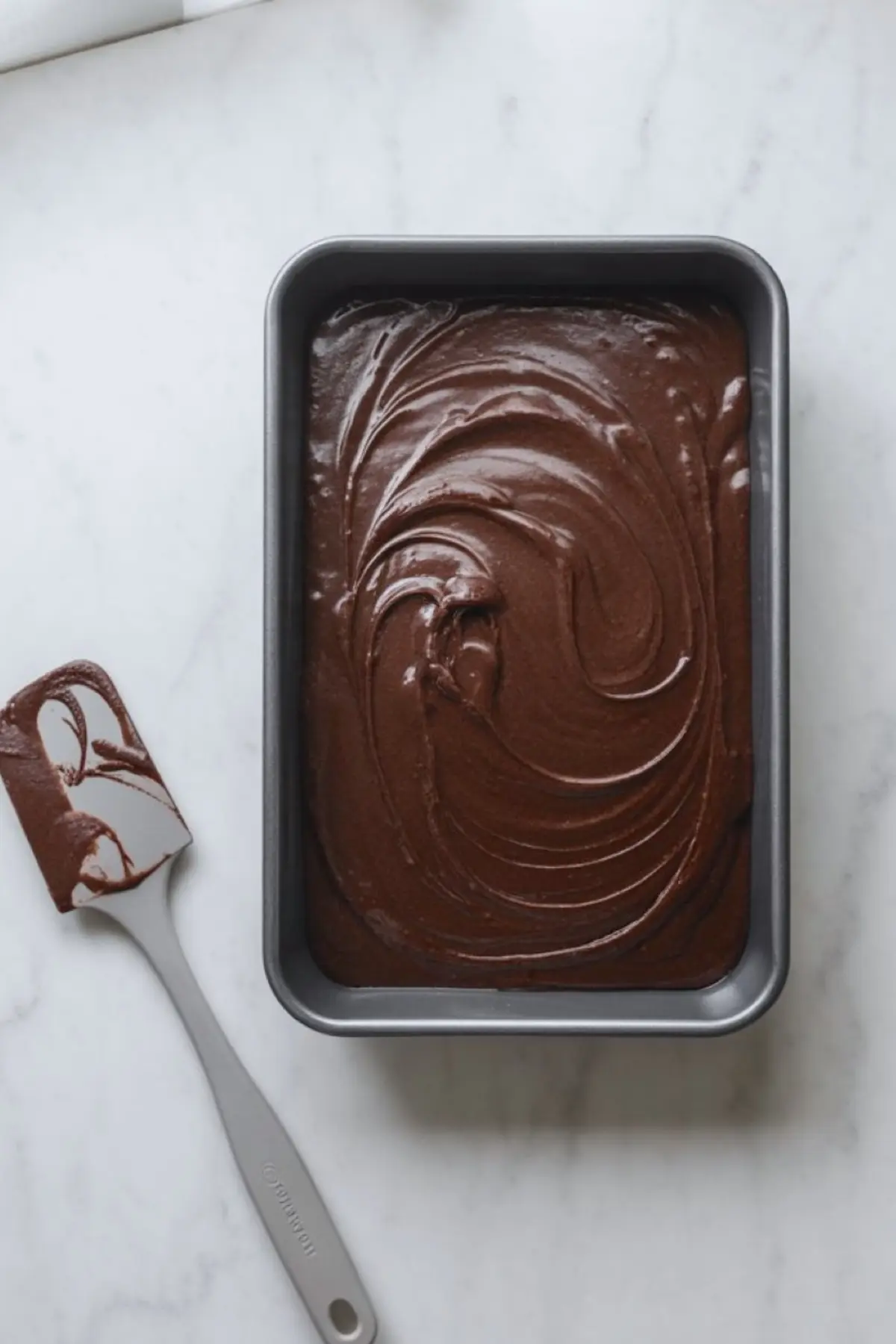 Chocolate cake batter fills a rectangular loaf pan on a marble countertop, and a silicone spatula rests beside the pan. The batter shows smooth swirls and glossy texture, creating a rich homemade chocolate cake base ready for baking.