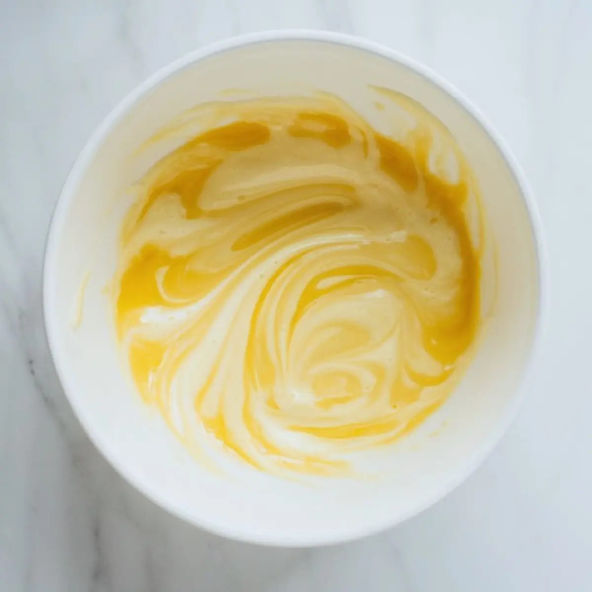 Egg and milk mixture swirls inside a white bowl on a marble countertop. The liquid shows yellow and creamy streaks, creating a smooth base for chocolate tres leches cake batter.