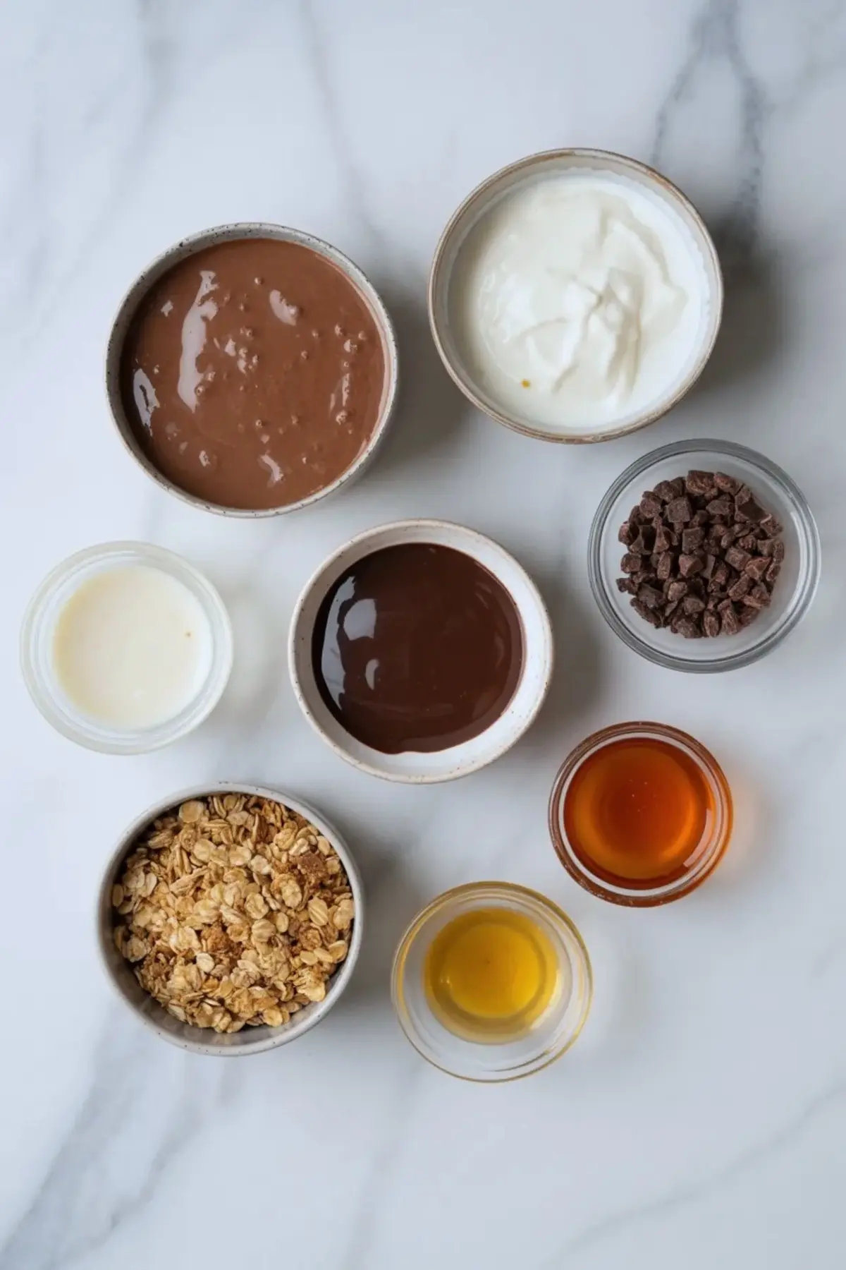 Ingredients for chocolate yogurt parfait arranged in bowls with yogurt, chocolate mixture, granola, cacao nibs, honey, and vanilla.