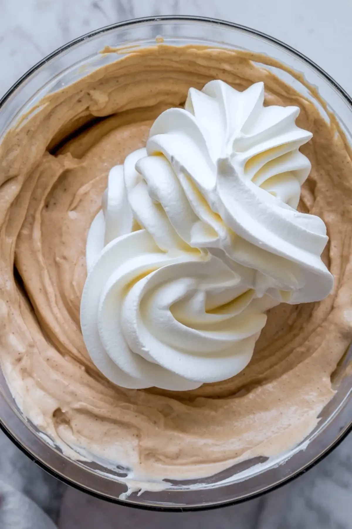 Glass bowl holds light brown coffee mousse mixture with a swirl of whipped cream on top during preparation of cappuccino coffee cheesecake filling.
