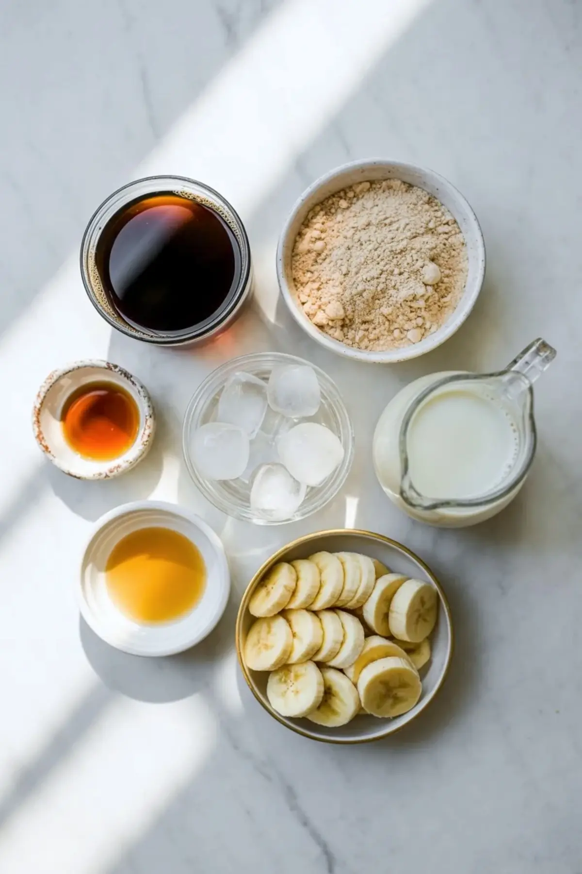 Overhead view shows coffee protein shake ingredients on white marble surface with brewed coffee, vanilla extract, maple syrup, banana slices, ice cubes, milk, and vanilla protein powder in small bowls.
