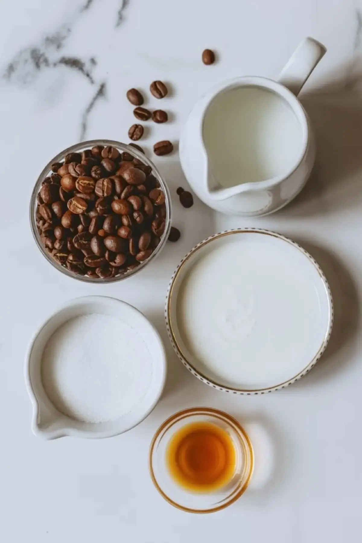 Coffee punch ingredients sit on a marble surface with coffee beans, cream, sugar, and vanilla measured in small bowls and pitchers.