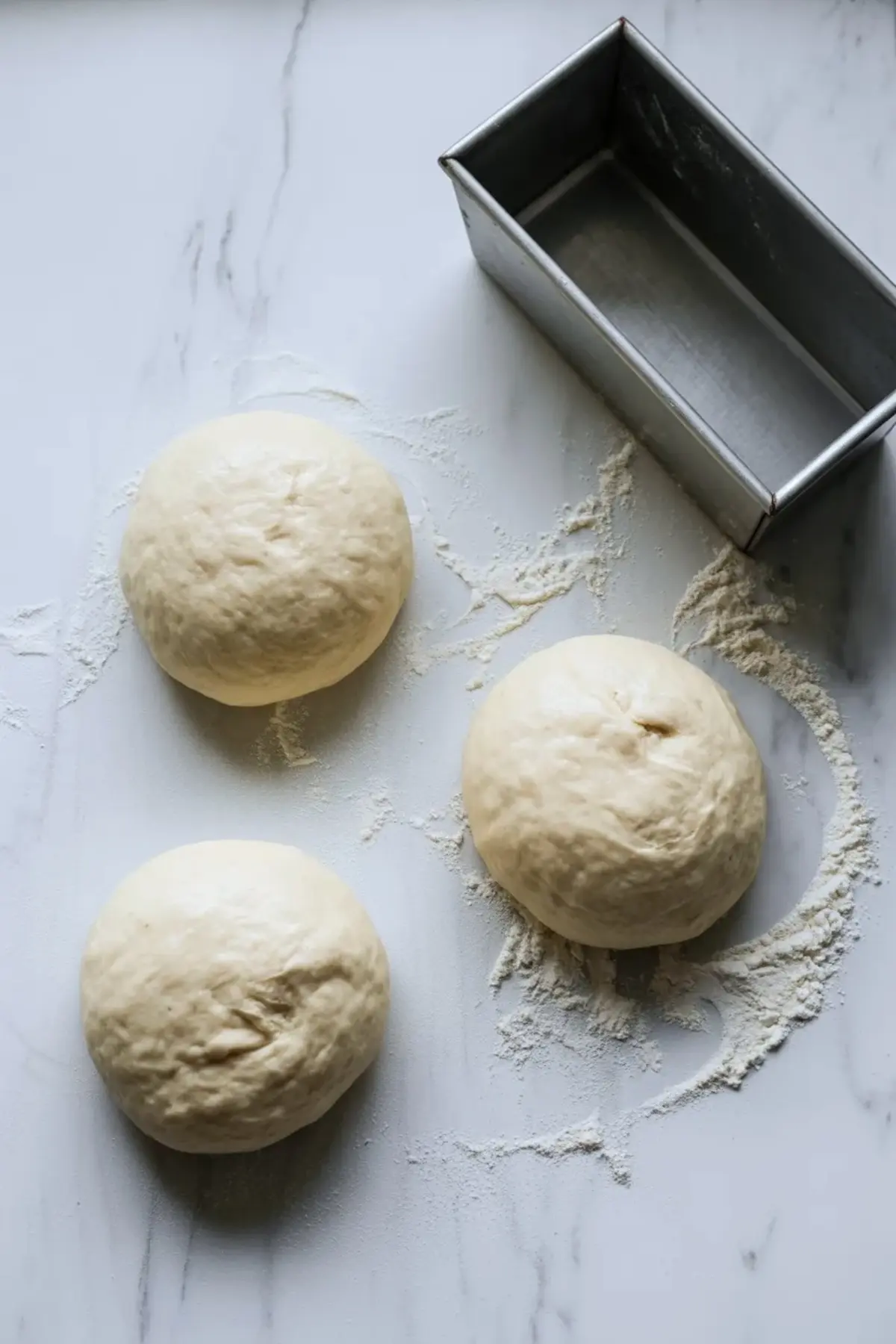 Three dough balls for sweet condensed milk bread beside a loaf pan on a floured marble surface.