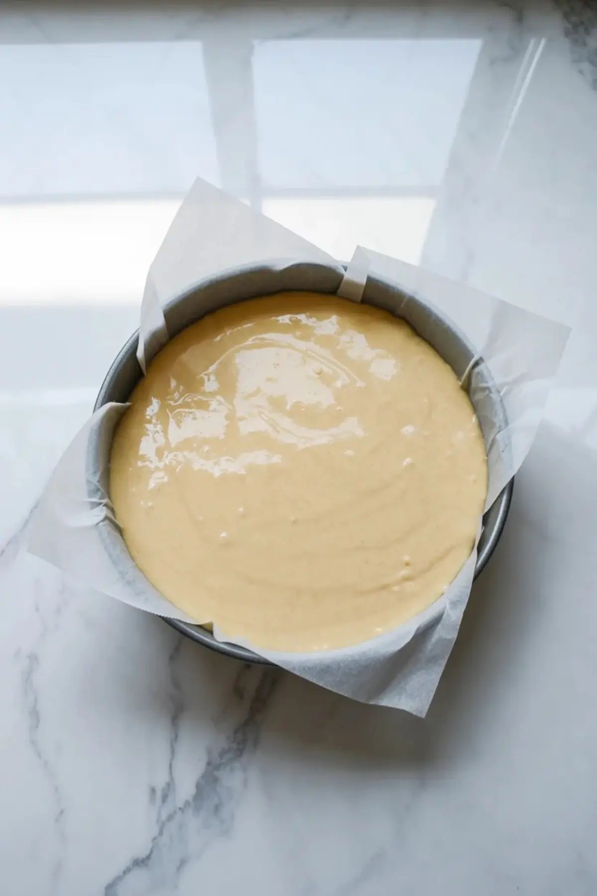 Round cake pan holds smooth pale cake batter lined with parchment paper on a white marble counter during condensed milk cake baking preparation.