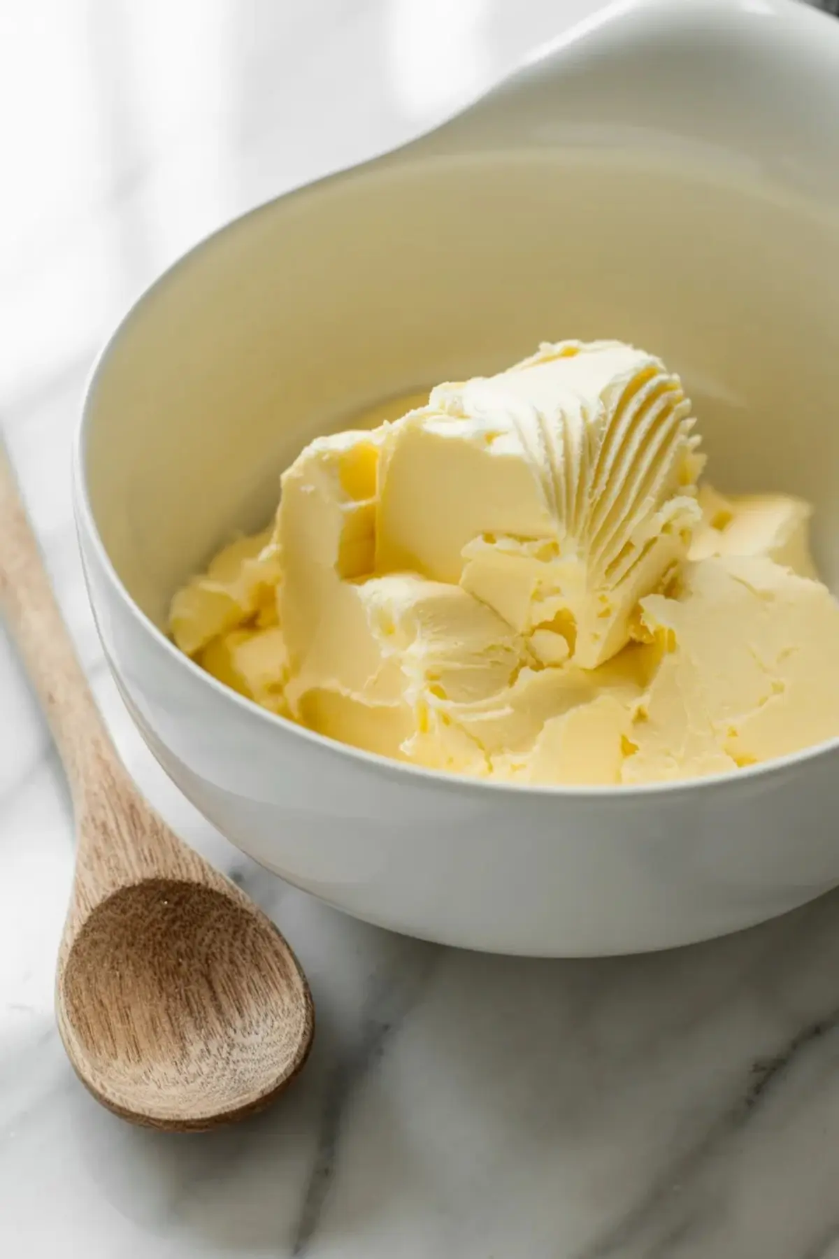 White bowl holds softened butter with ridged texture beside a wooden spoon on a marble countertop for homemade cake recipe preparation.