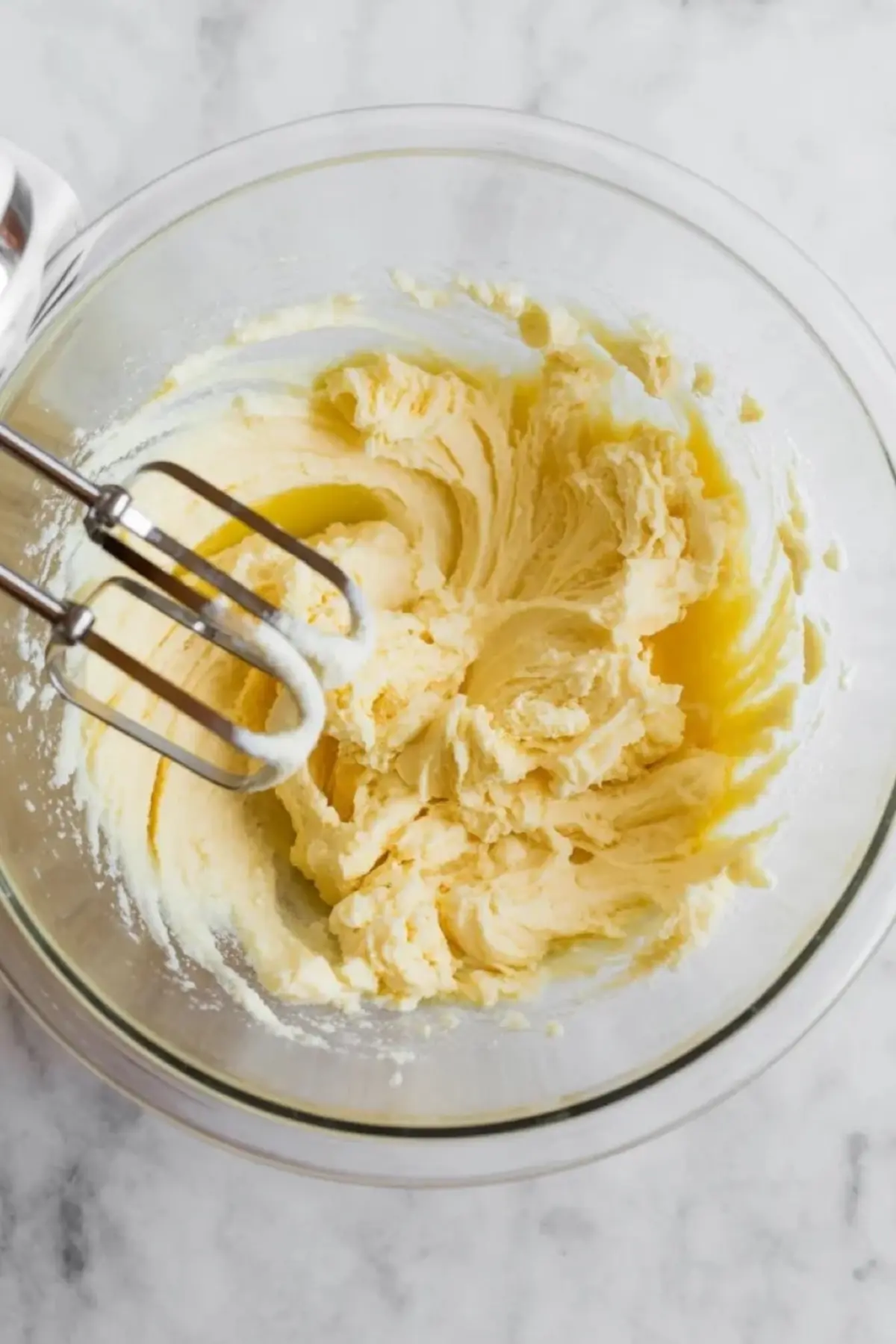 A glass mixing bowl holds creamed butter and sugar blended into a smooth pale mixture. An electric hand mixer blends the soft dough base for condensed milk cookies.