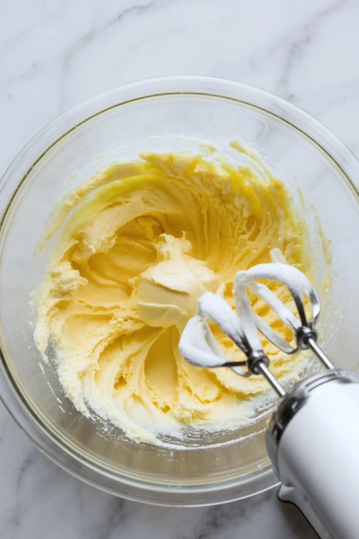 Hand mixer beats butter and sugar in a glass mixing bowl to create a light fluffy buttercream base for Fruity Pebbles frosting on a white marble countertop.
