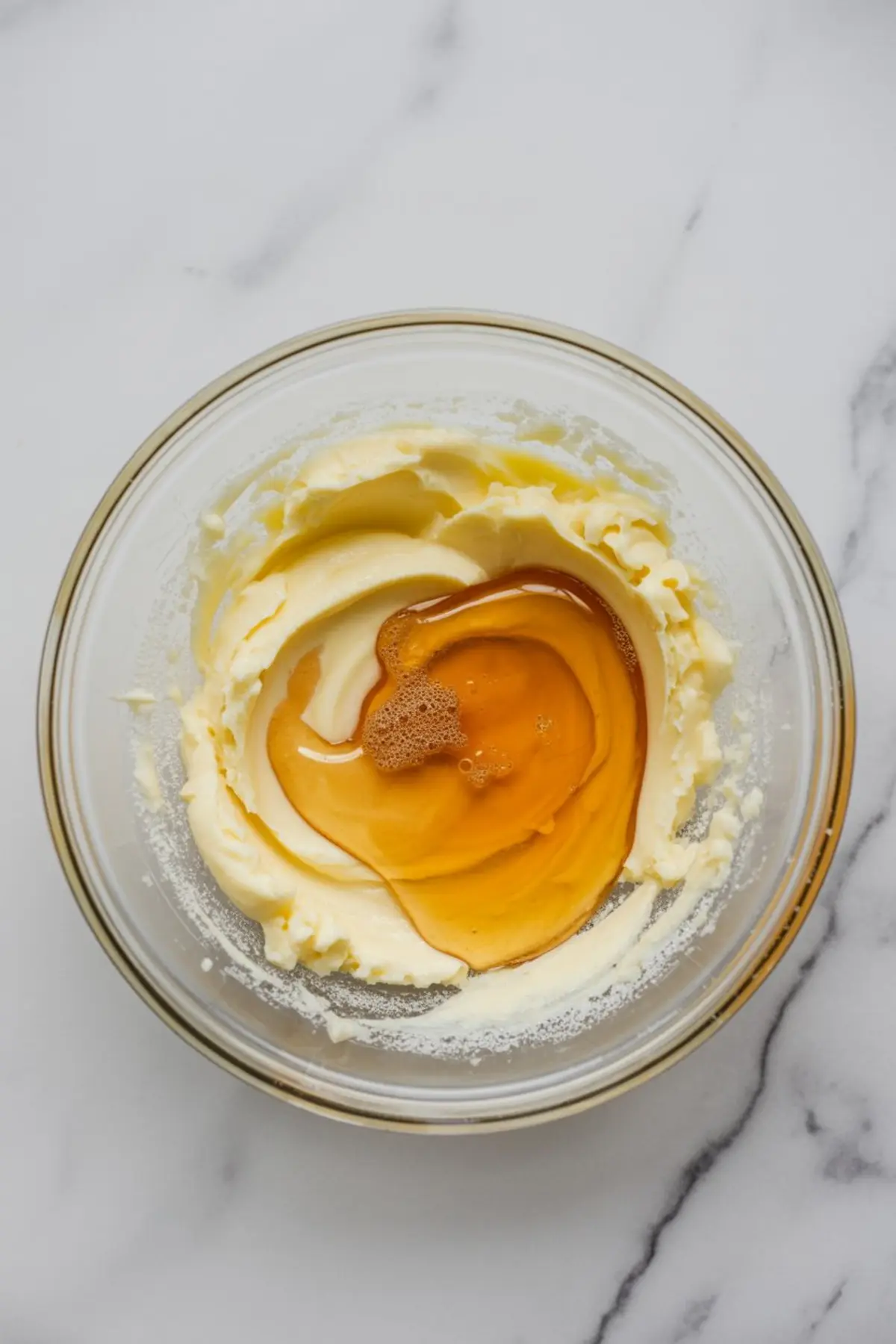 Glass bowl holds softened butter and golden honey on a white marble surface. Honey pools over the butter in a thick swirl, showing the start of homemade honey frosting or honey buttercream recipe.
