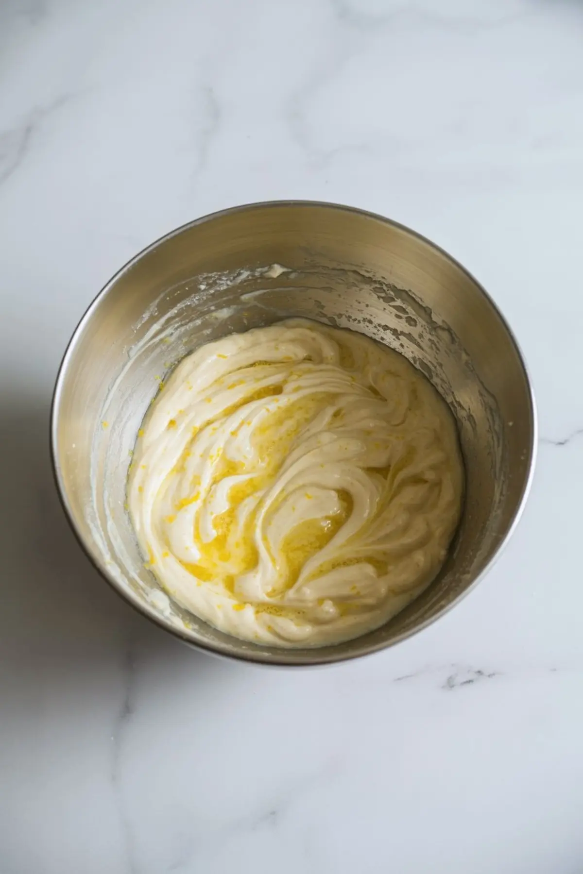 Metal mixing bowl holds lemon cake batter with yellow lemon juice streaks on a white marble surface during lemon pudding cake recipe preparation.
