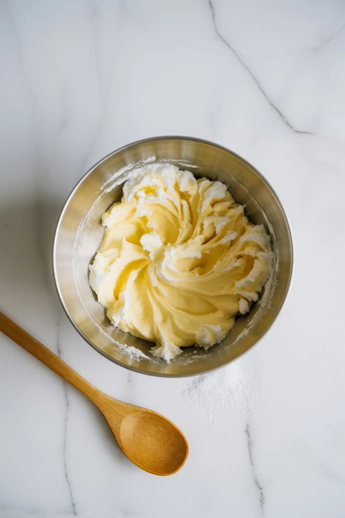 Metal bowl holds creamed butter and sugar mixture with wooden spoon and powdered sugar on white marble surface during lemon pudding cake batter preparation.

