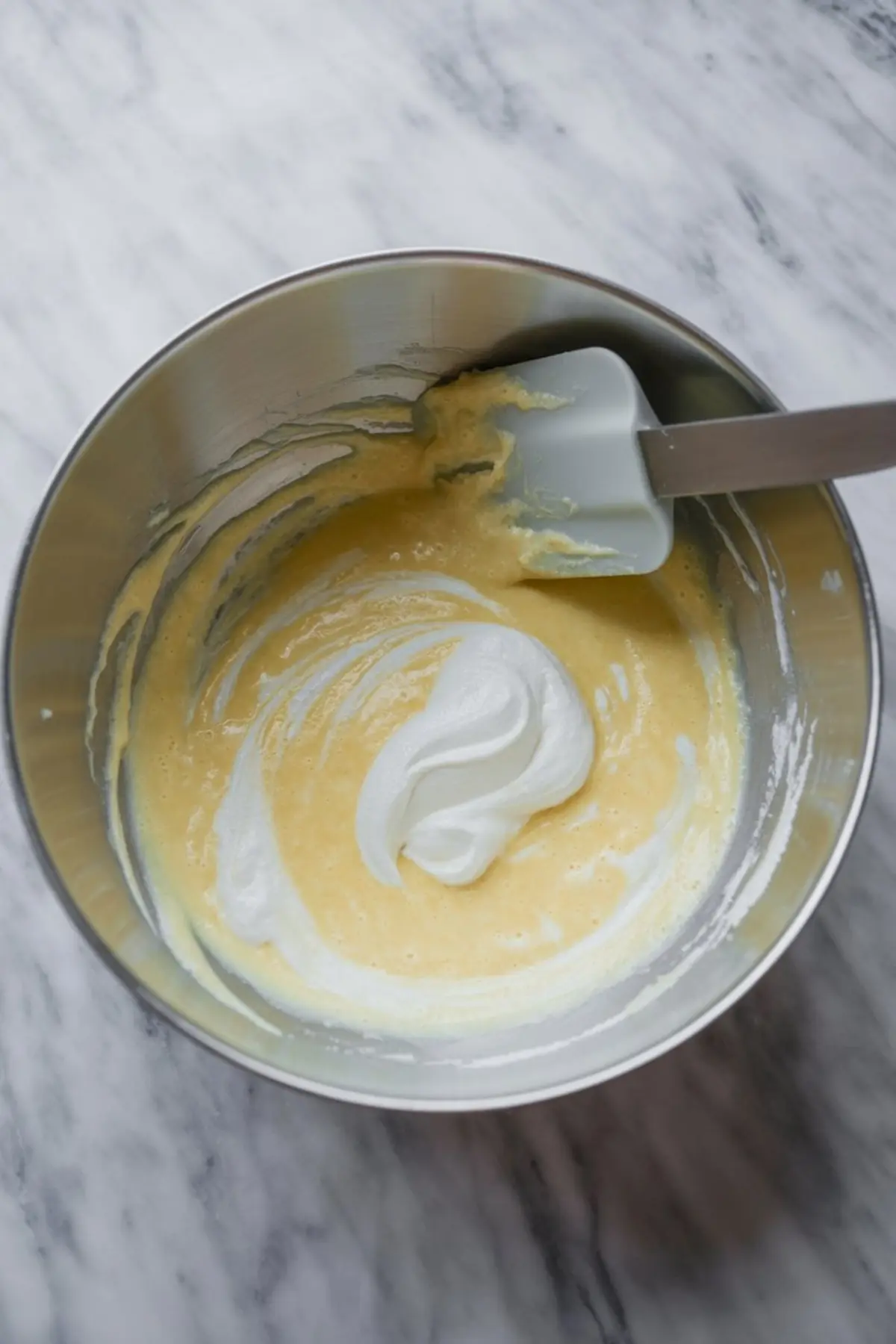 Metal mixing bowl shows lemon cake batter with folded whipped egg whites and silicone spatula during light lemon pudding cake batter mixing step.
