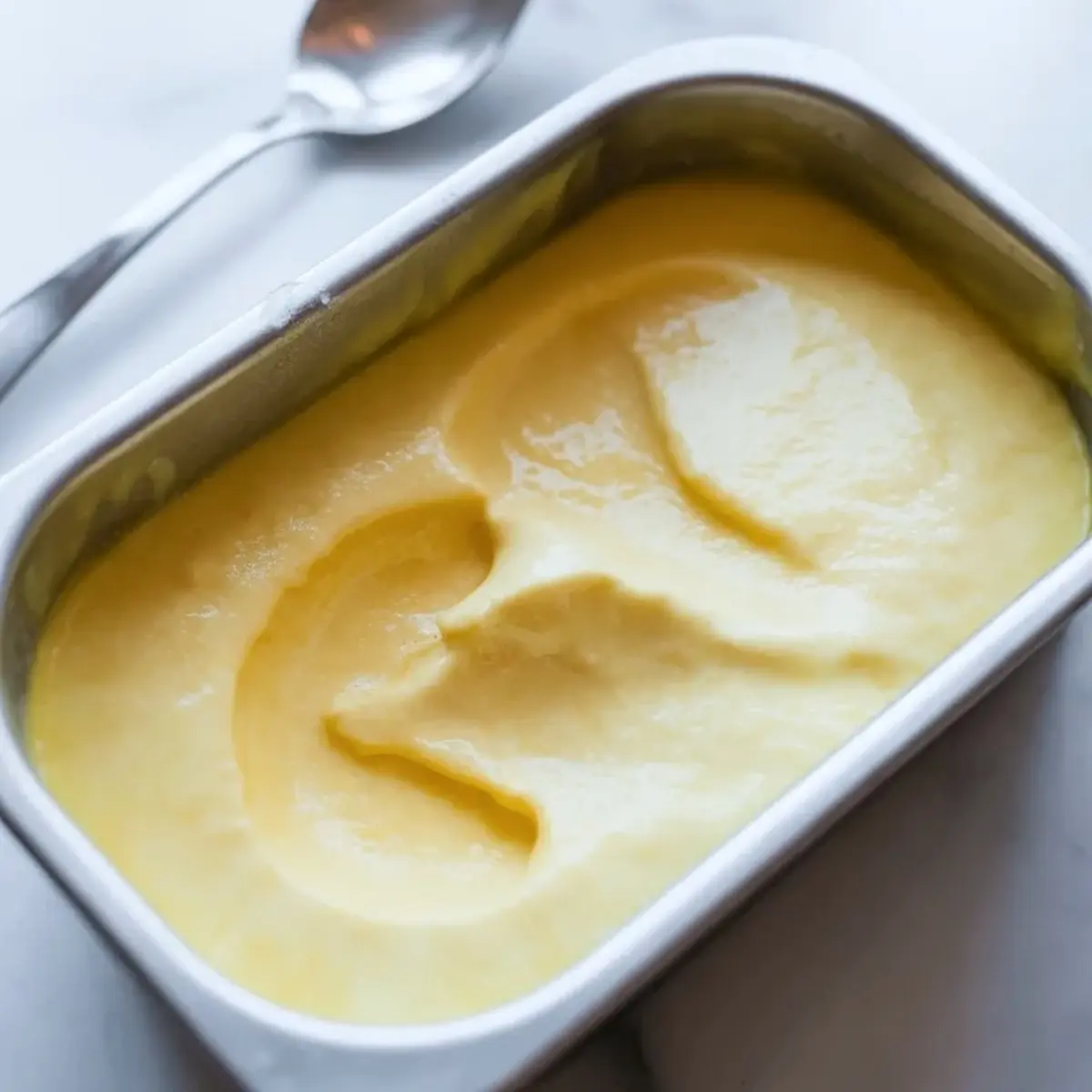 Fresh lemon sorbet with smooth swirls in a loaf pan on a white surface beside a spoon.